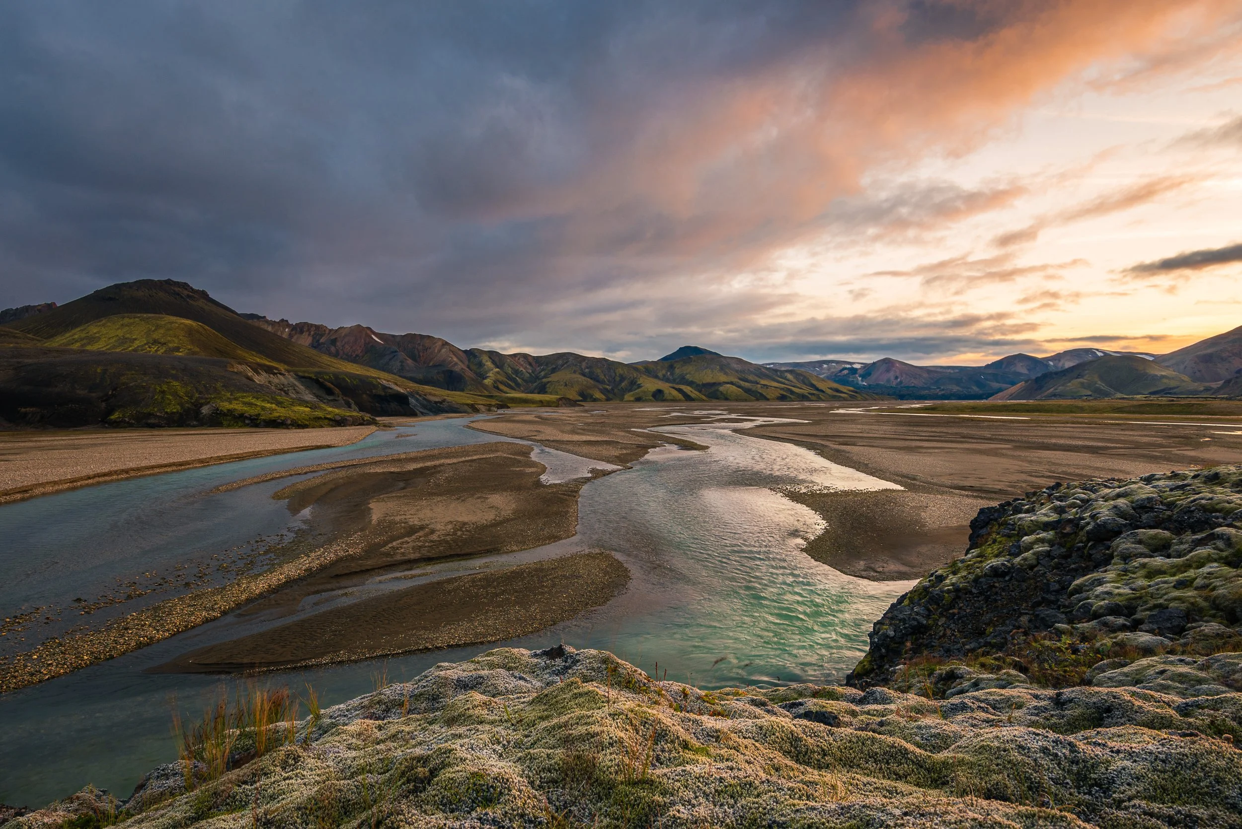 A winding river flowing through a wide valley with green mountains in the background during sunset.
