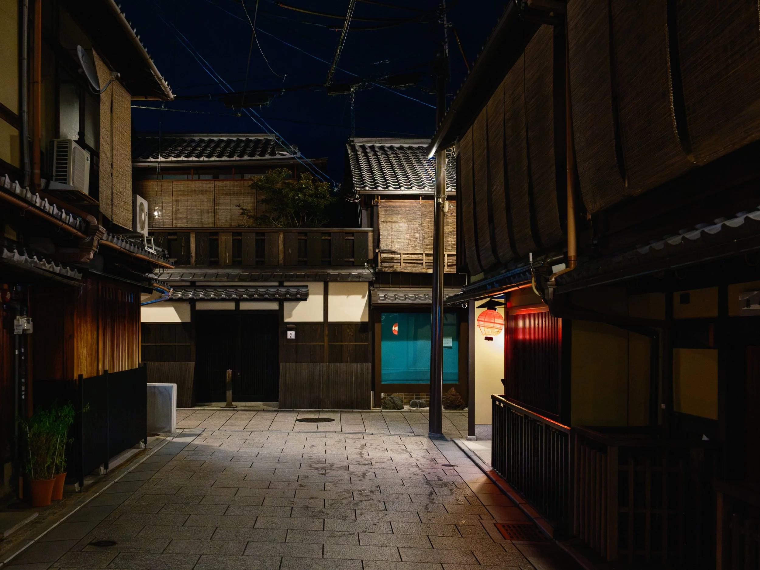 A quiet, illuminated alleyway at night, featuring traditional Japanese-style buildings with tiled roofs, wooden walls, and a hanging red paper lantern.