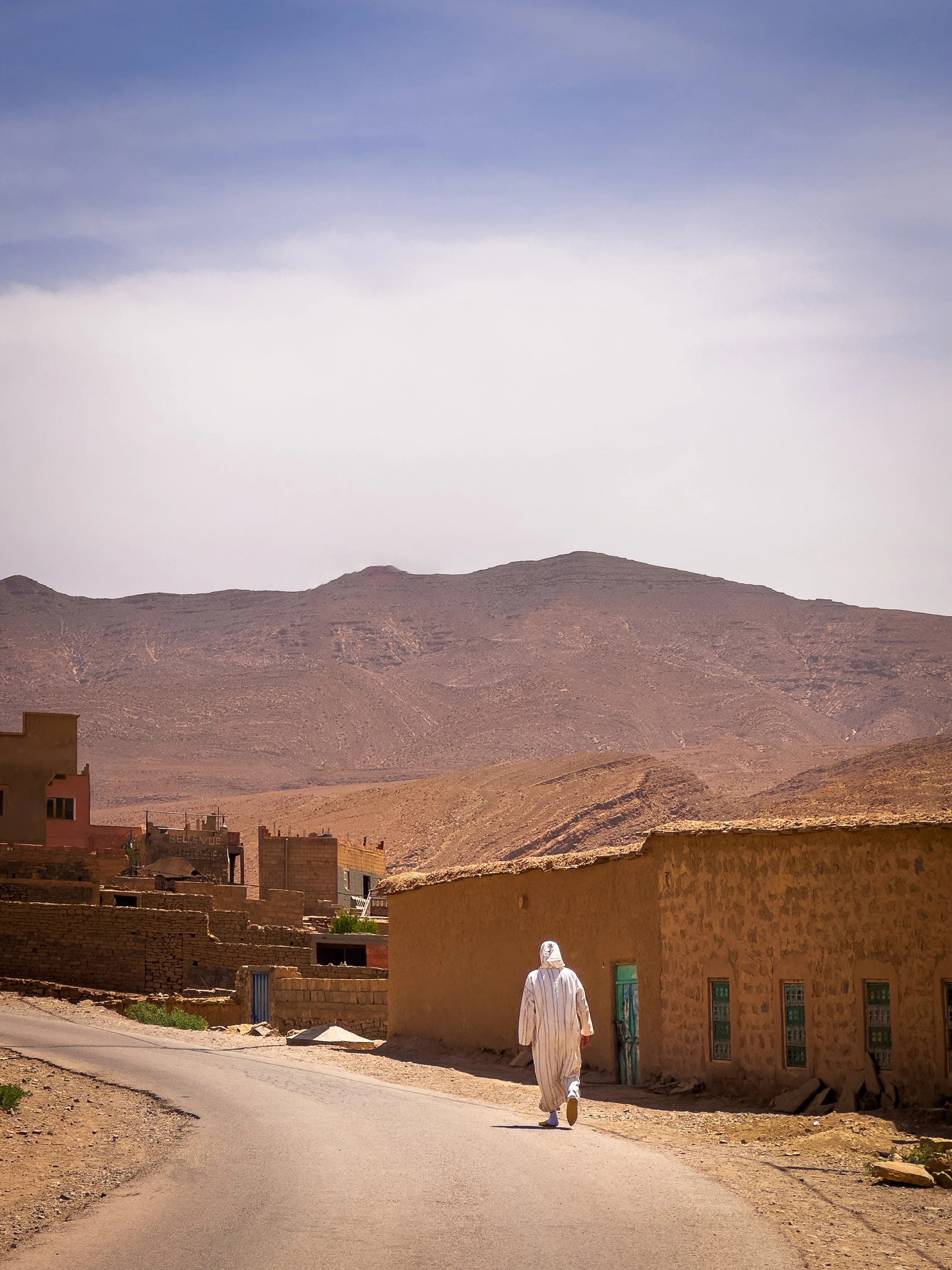 A person in traditional clothing walking along a dirt road in a desert landscape with small houses and mountains in the background.