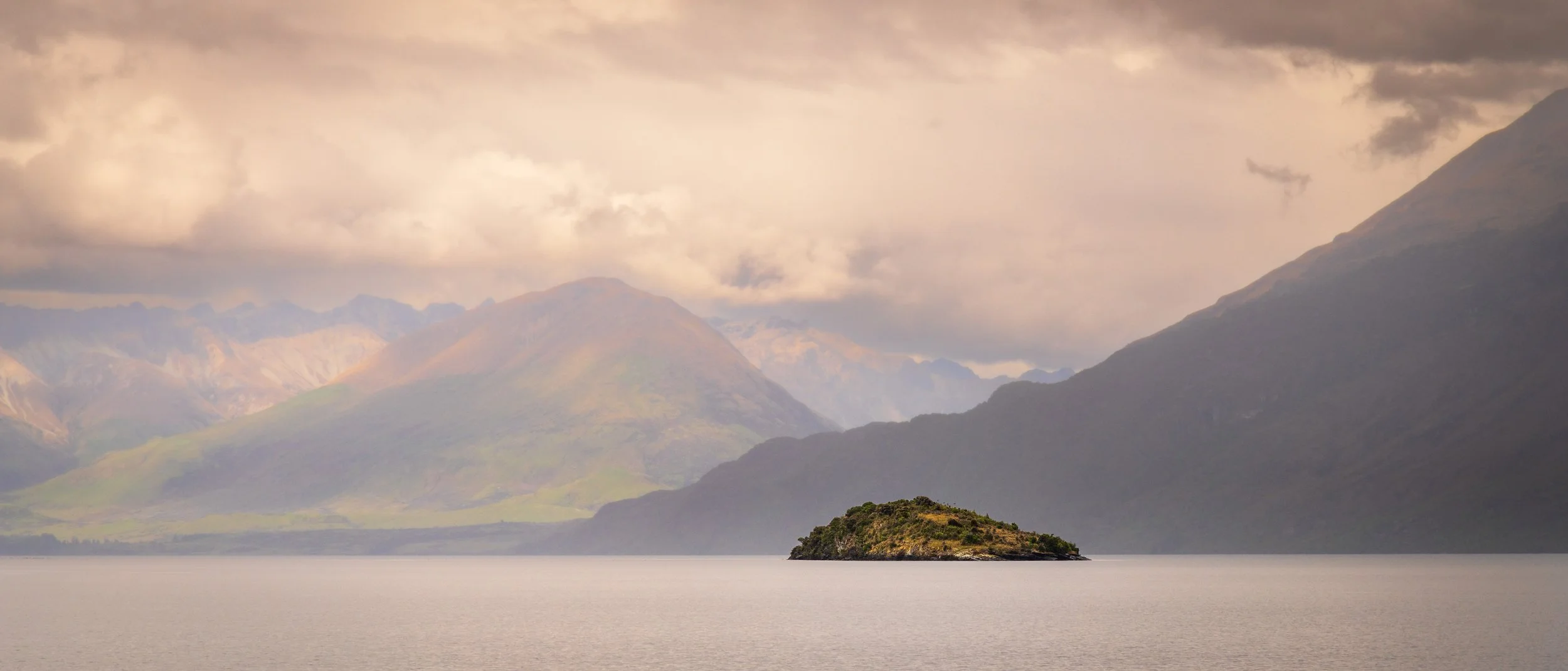 A small island with greenery in a calm body of water, surrounded by mountains under a cloudy sky.