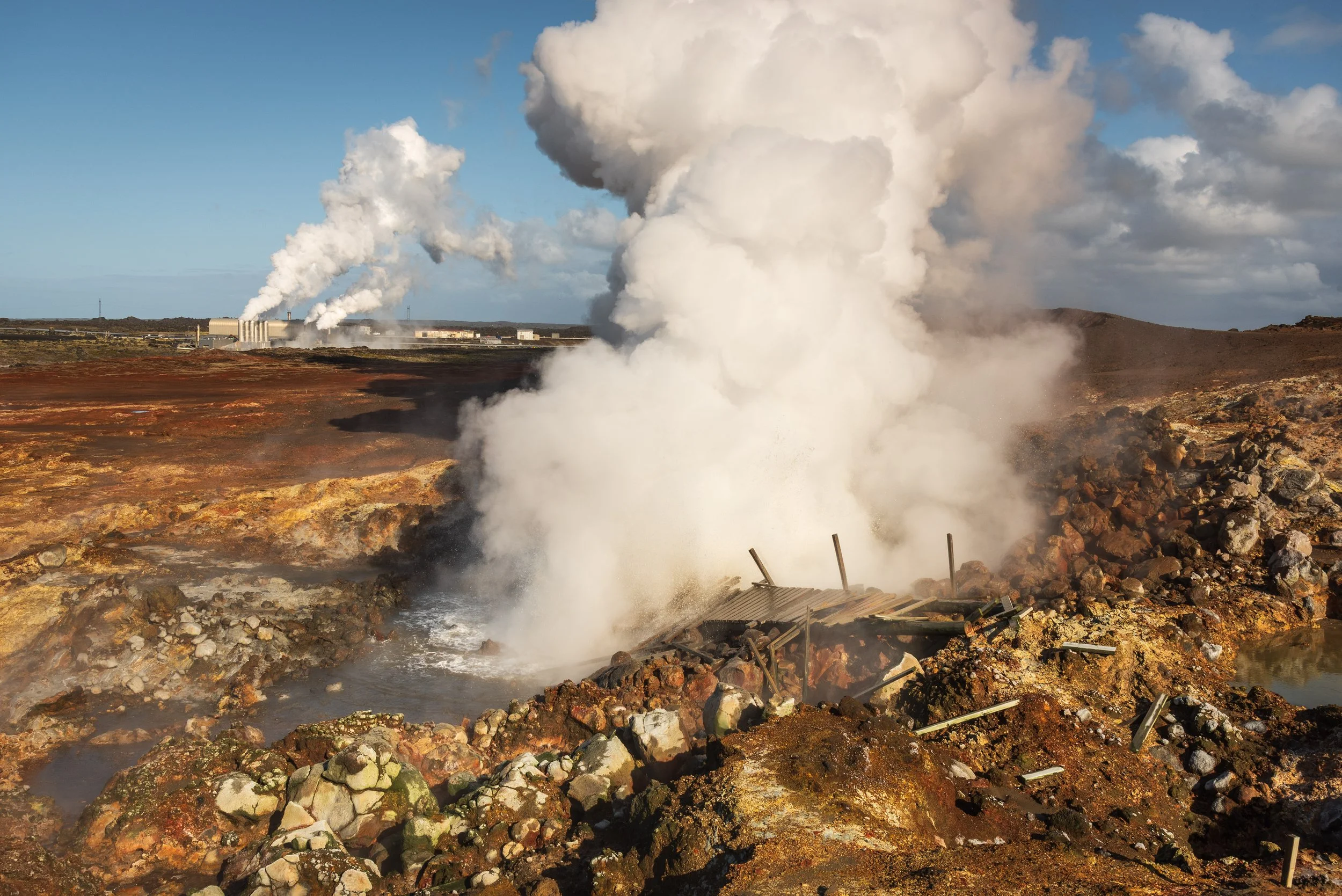 Geothermal scene with bubbling ground, steam venting, and industrial buildings in the background.