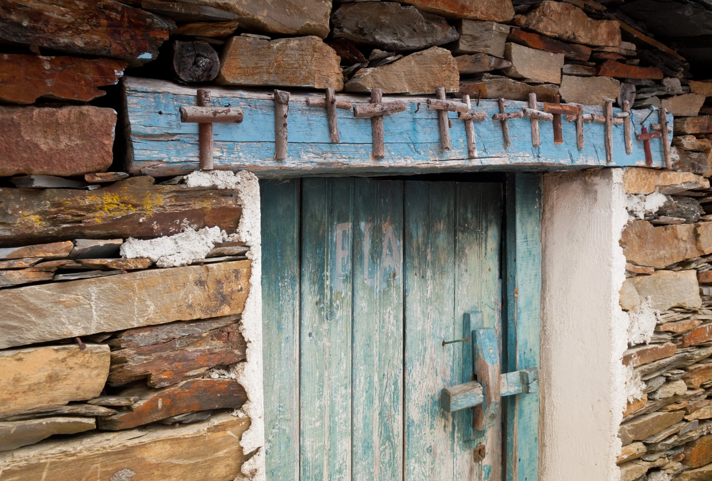 Old weathered wooden door with peeling paint and a makeshift latch, set within a stone wall with uneven rocks and a wooden lintel above.