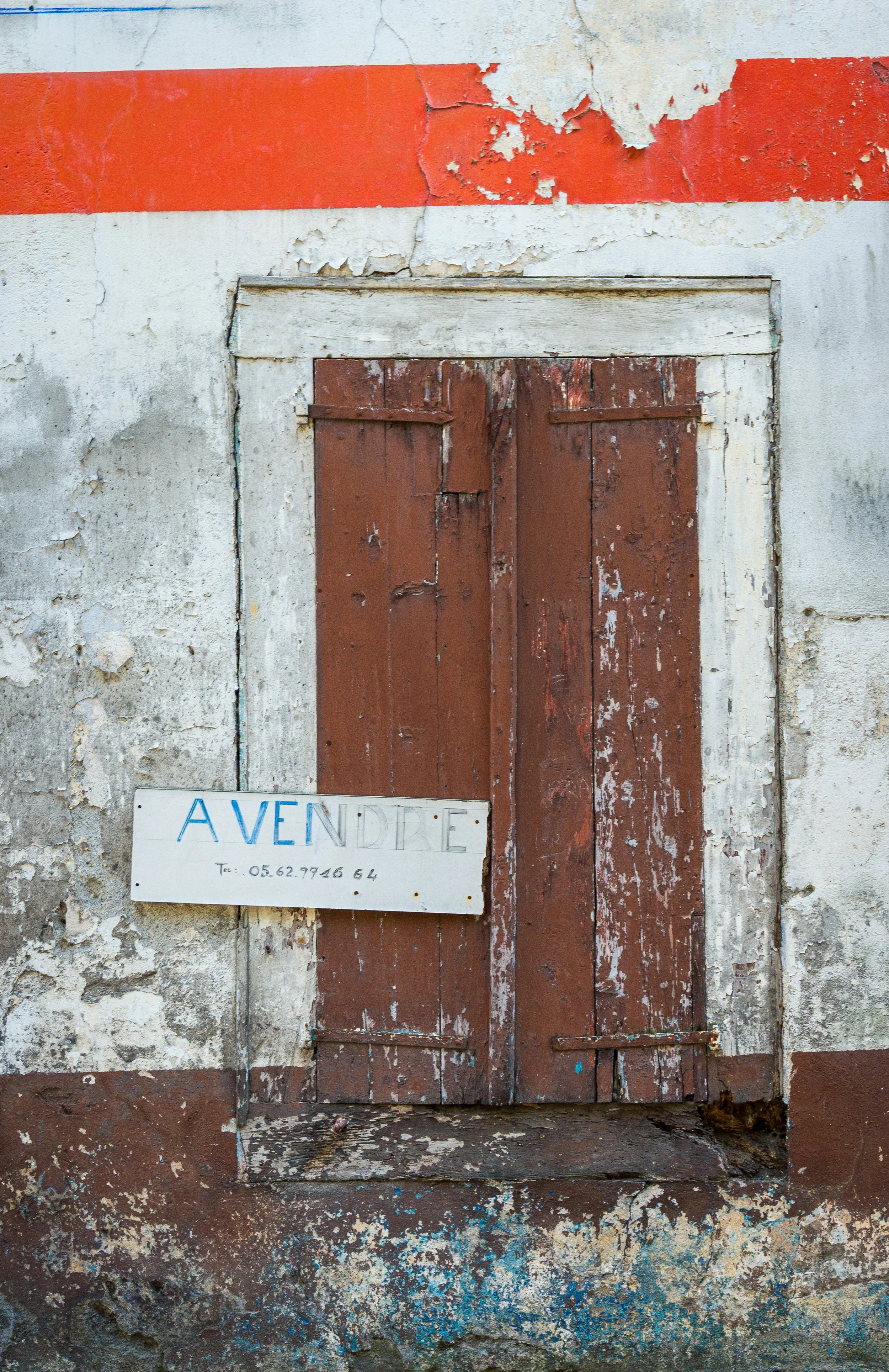 A weathered, red wooden door with peeling paint set in a decayed, concrete wall with cracks and peeling paint. A white sign with blue and black text reading 'AVEN DRE' and a phone number is leaning against the wall near the bottom left of the door.
