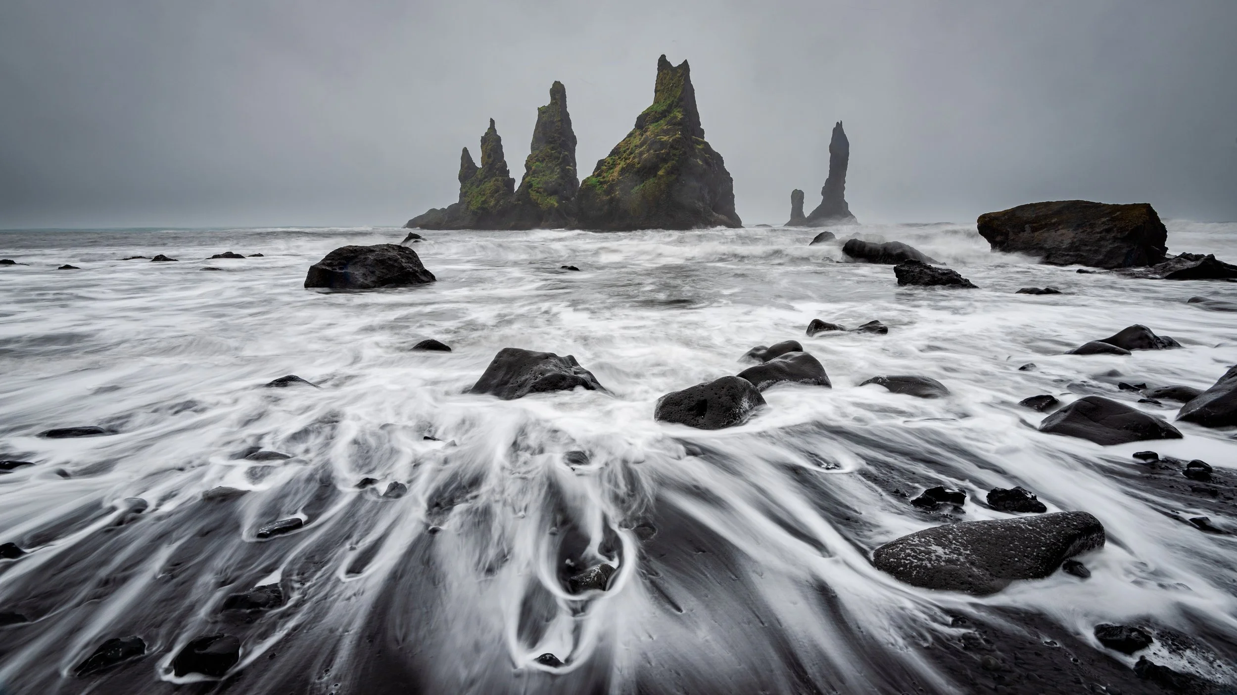 Rocky black sand beach with waves washing over rocks and sea stacks in the distance under a cloudy sky.