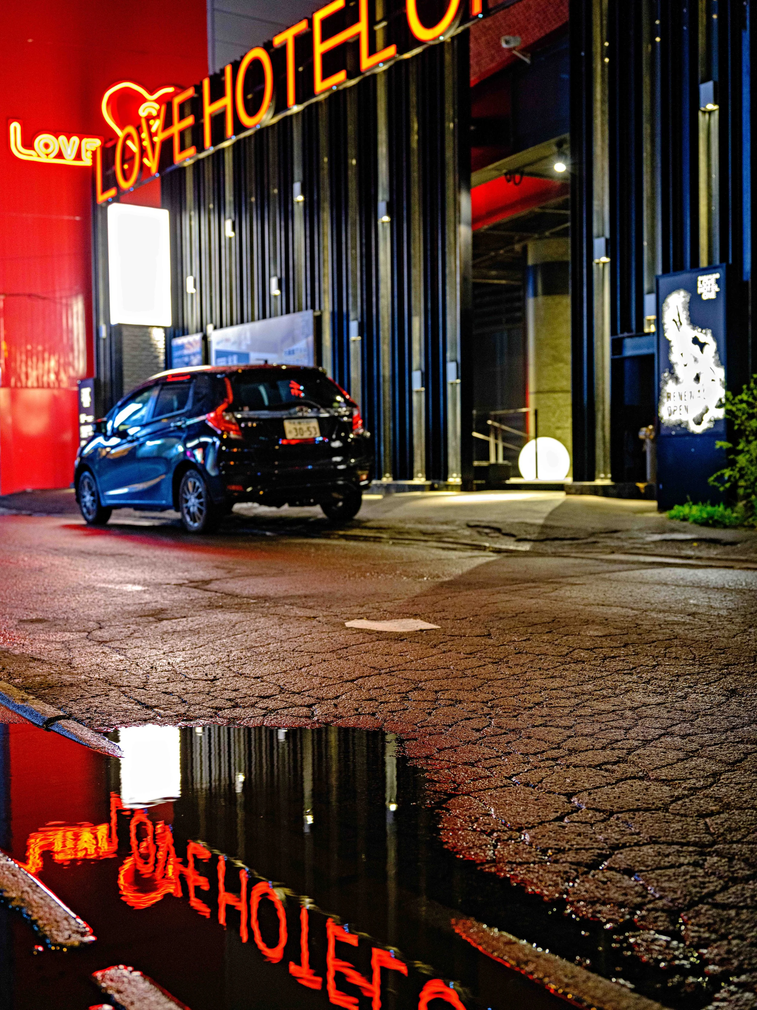 Night scene outside a building with neon signs reading 'LOVE' and 'LOVE HOTEL', a parked black SUV, and a puddle reflecting the red neon signs.