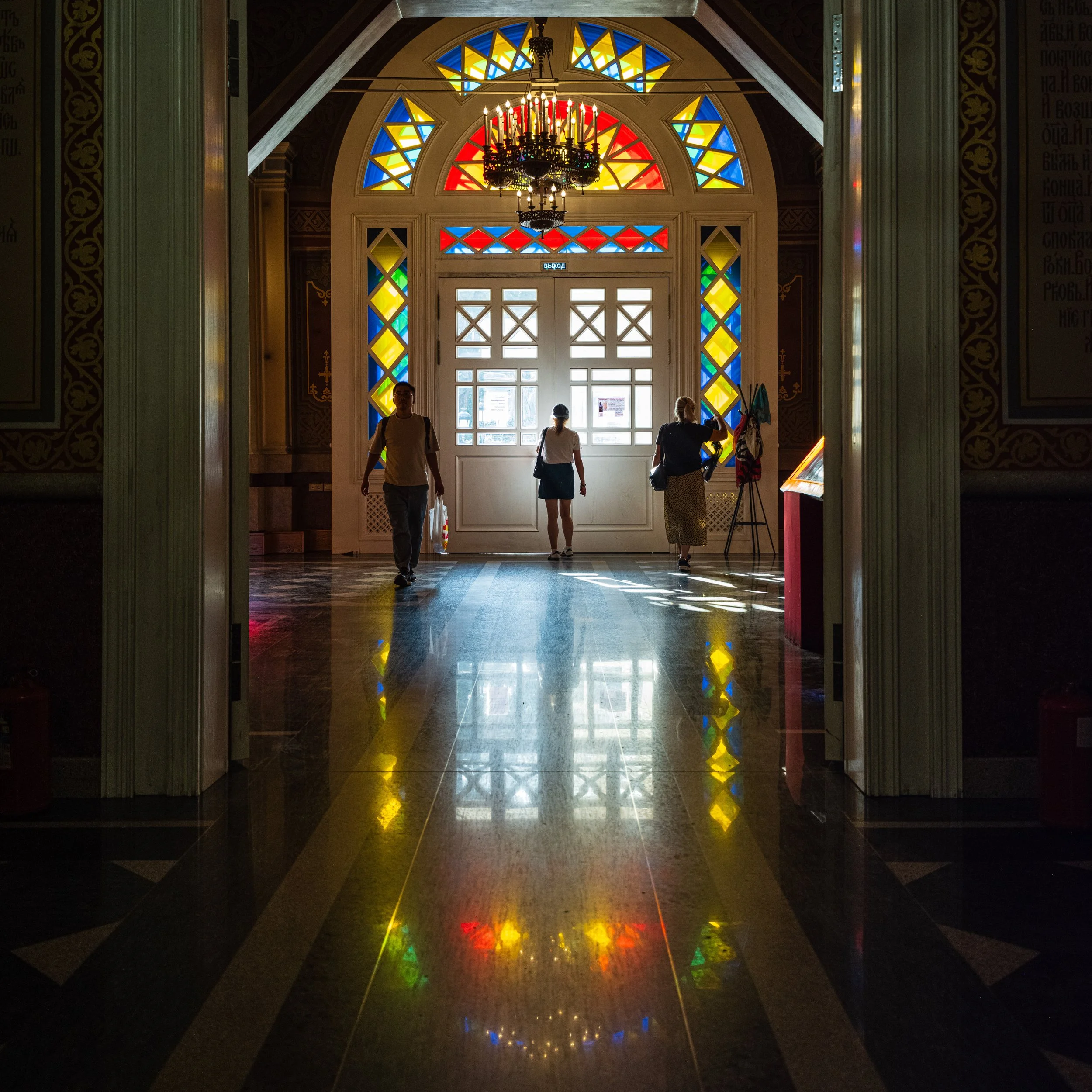 Interior view of a building with a colorful stained glass window door and a chandelier, with three people entering or exiting, their silhouettes visible against the bright light outside.