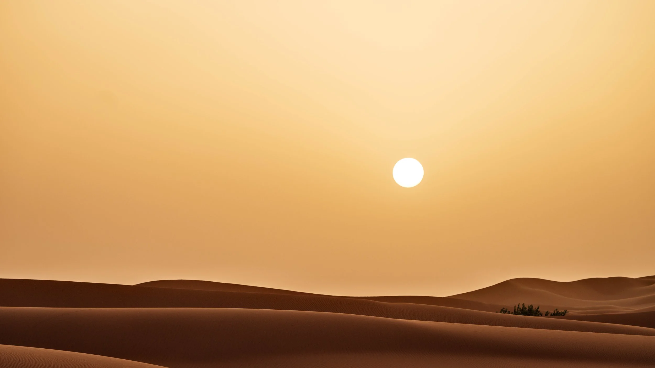 A desert landscape at sunset with sand dunes and a bright sun in the sky.