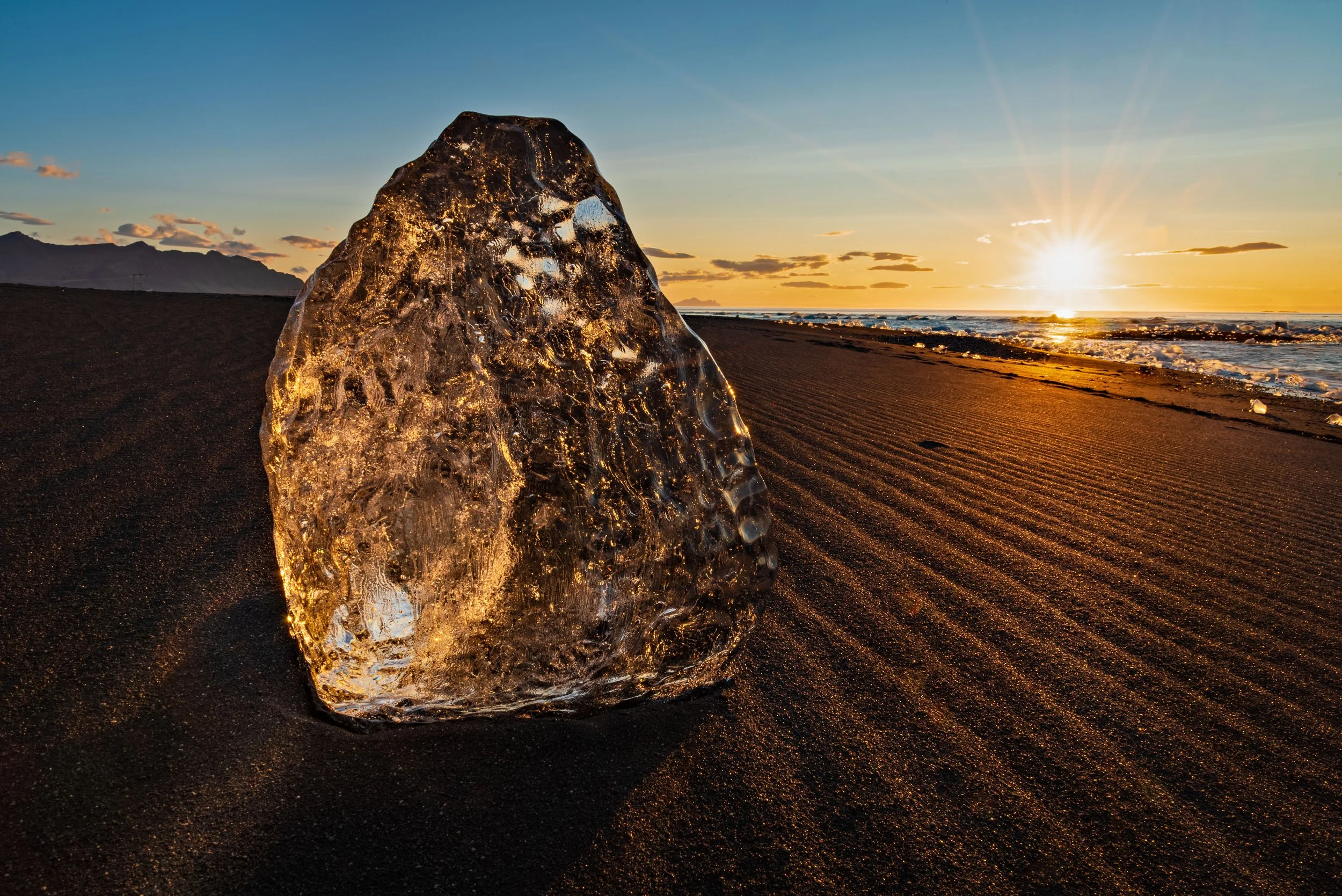 A large, clear ice chunk on dark sandy beach at sunset, with the sun low on the horizon over the ocean, creating a warm glow and reflected light on the ice.