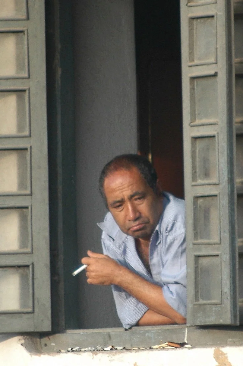A man leaning out of a window, holding a cigarette, and looking pensive