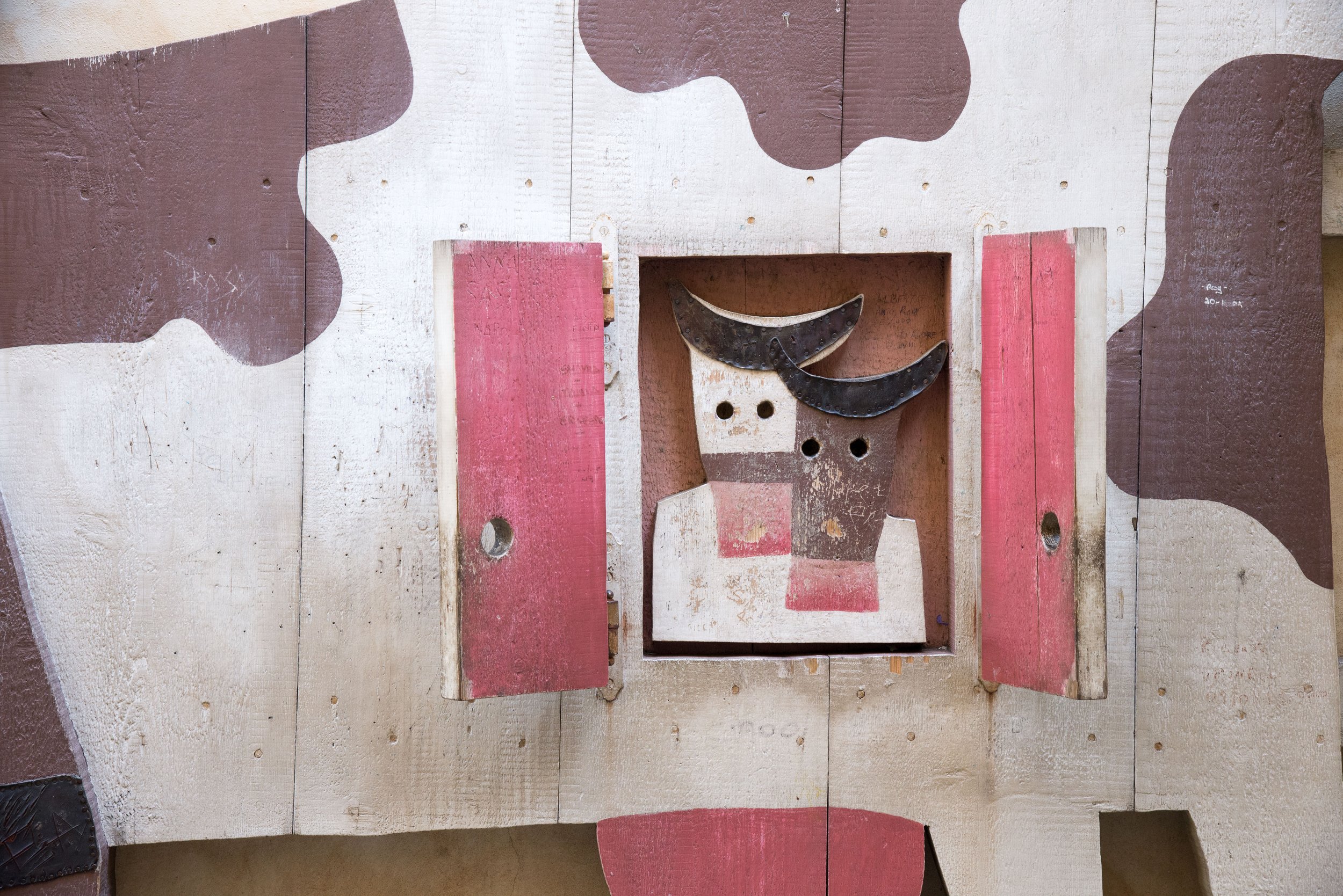 Wooden artwork of a cow's face with two black horns, set in a frame with a red and white background, surrounded by painted brown and white abstract shapes on a wooden wall.