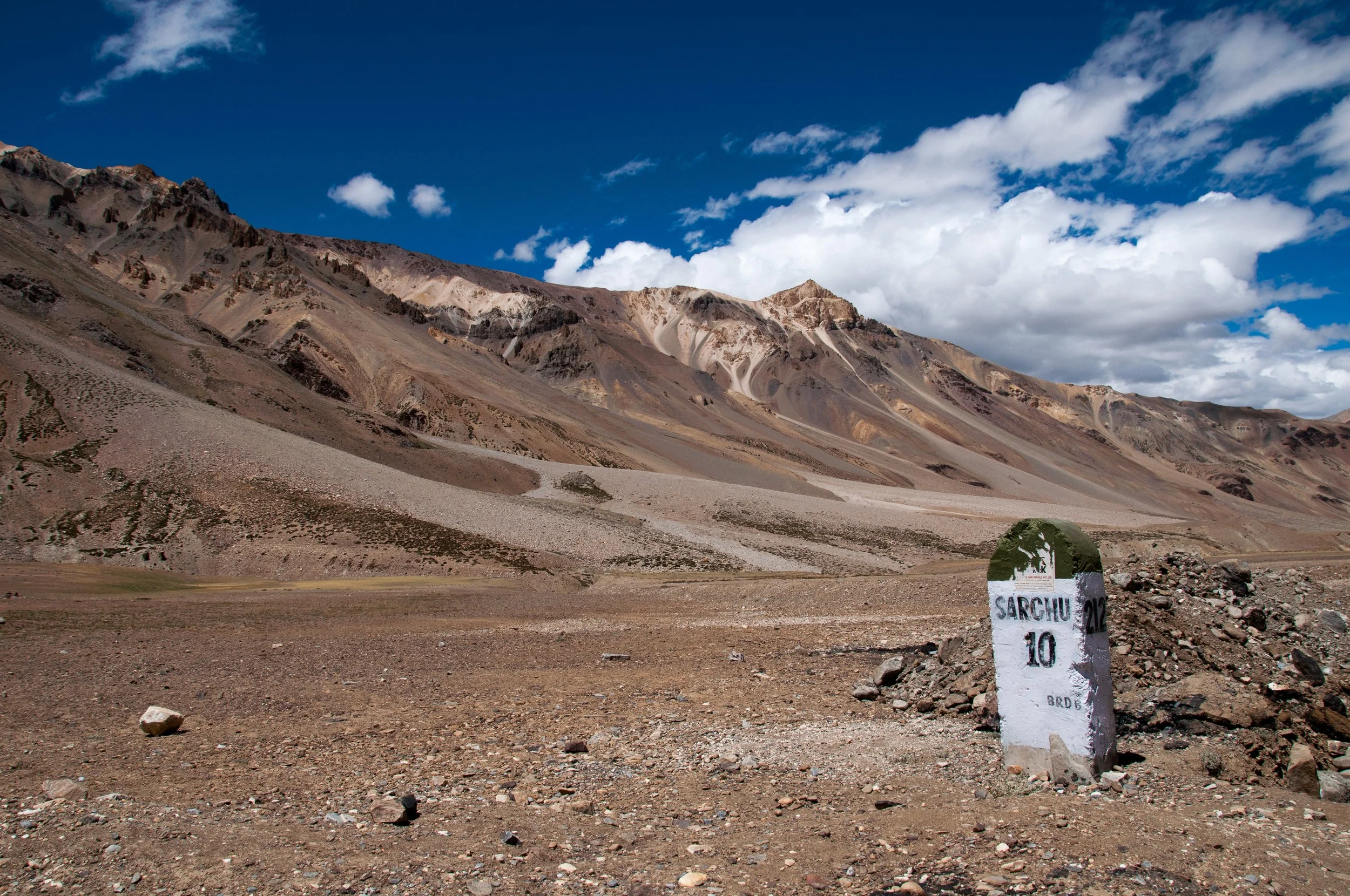 A barren mountain landscape with a clear blue sky and white clouds, featuring a stone milestone marked 'Sarchu 10' in the foreground.