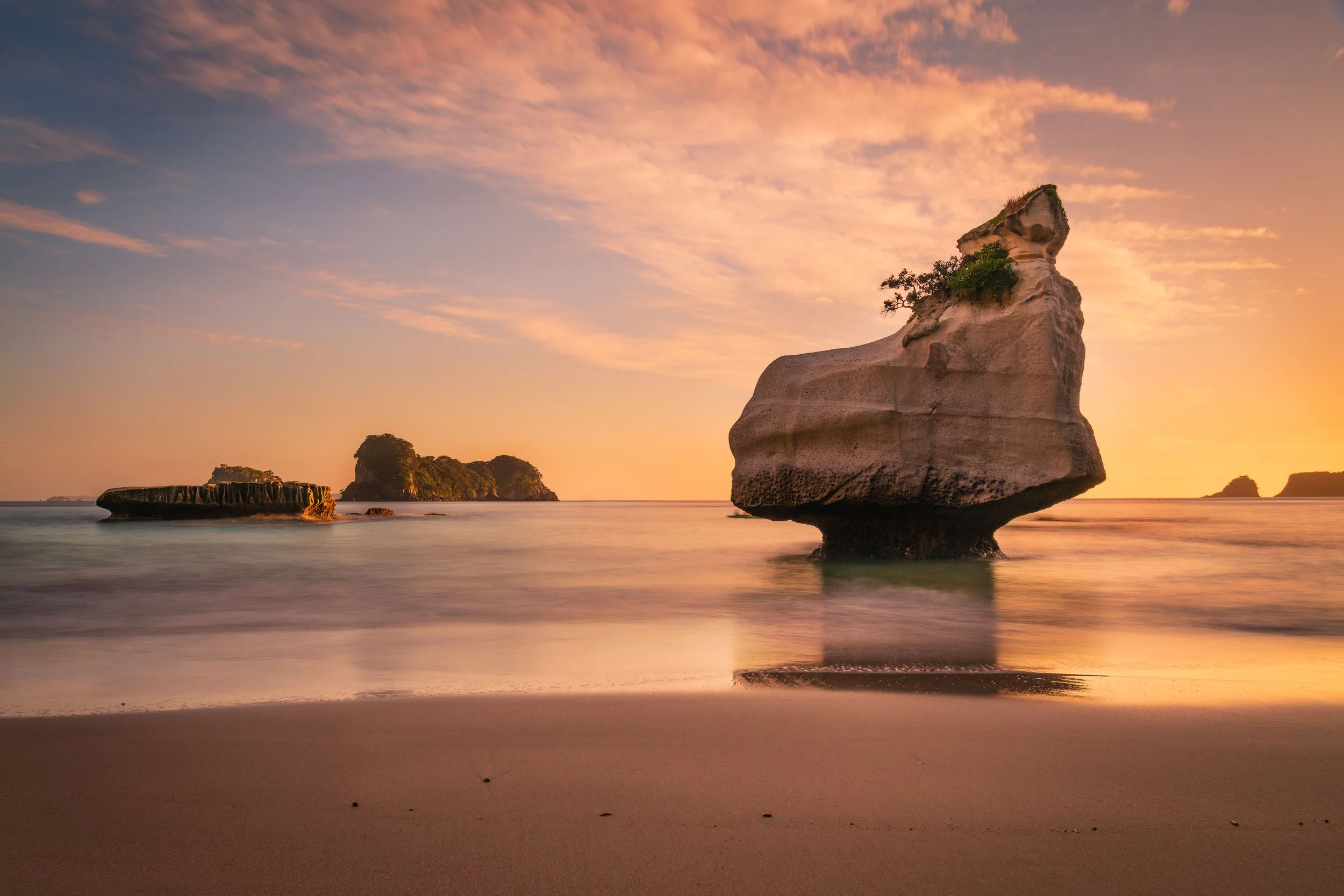 Seashore during sunset with large rock formations in calm water and a pastel orange sky with wispy clouds.