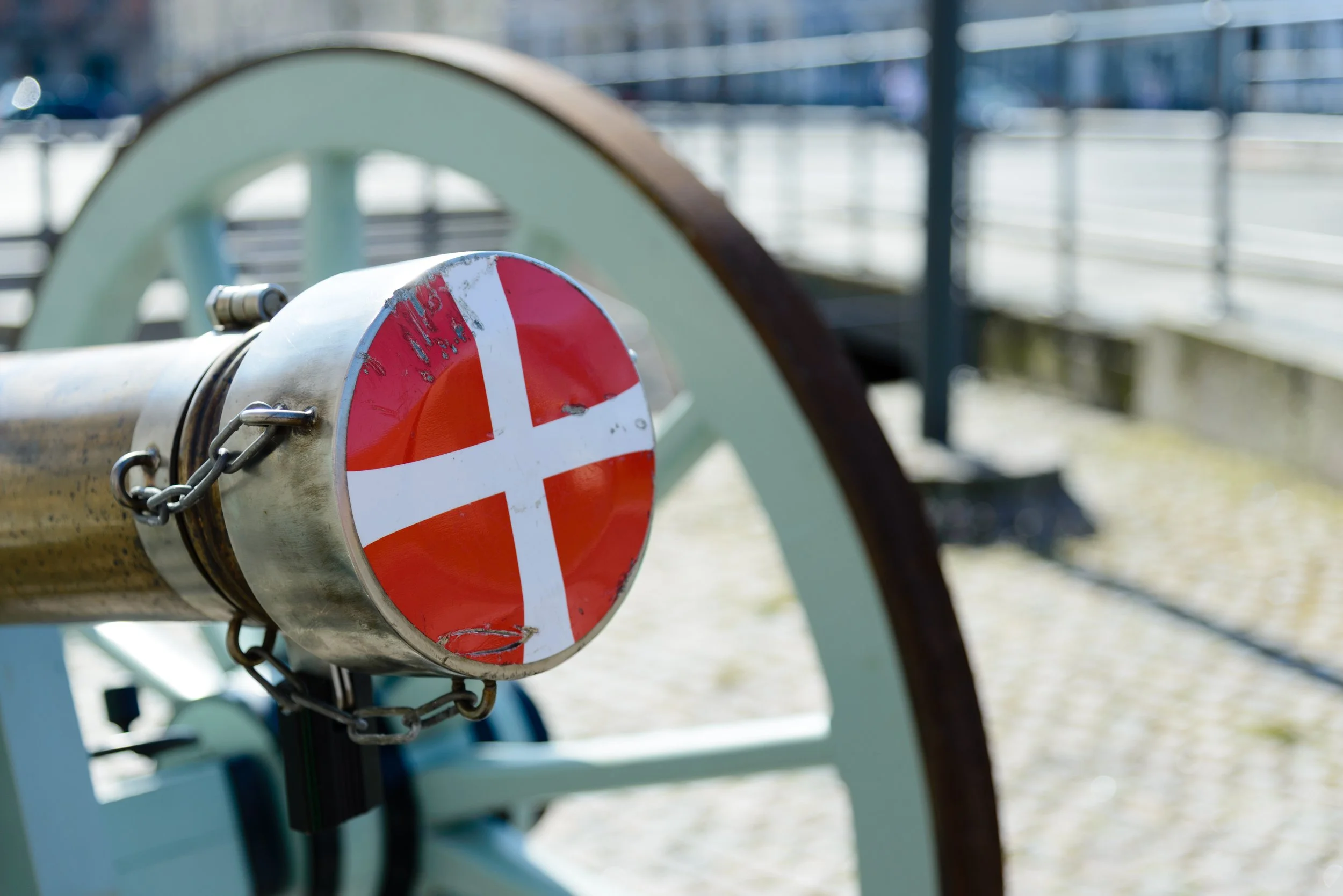 Close-up of a vintage cannons with a Danish flag sticker on the barrel, outdoors near a waterfront.