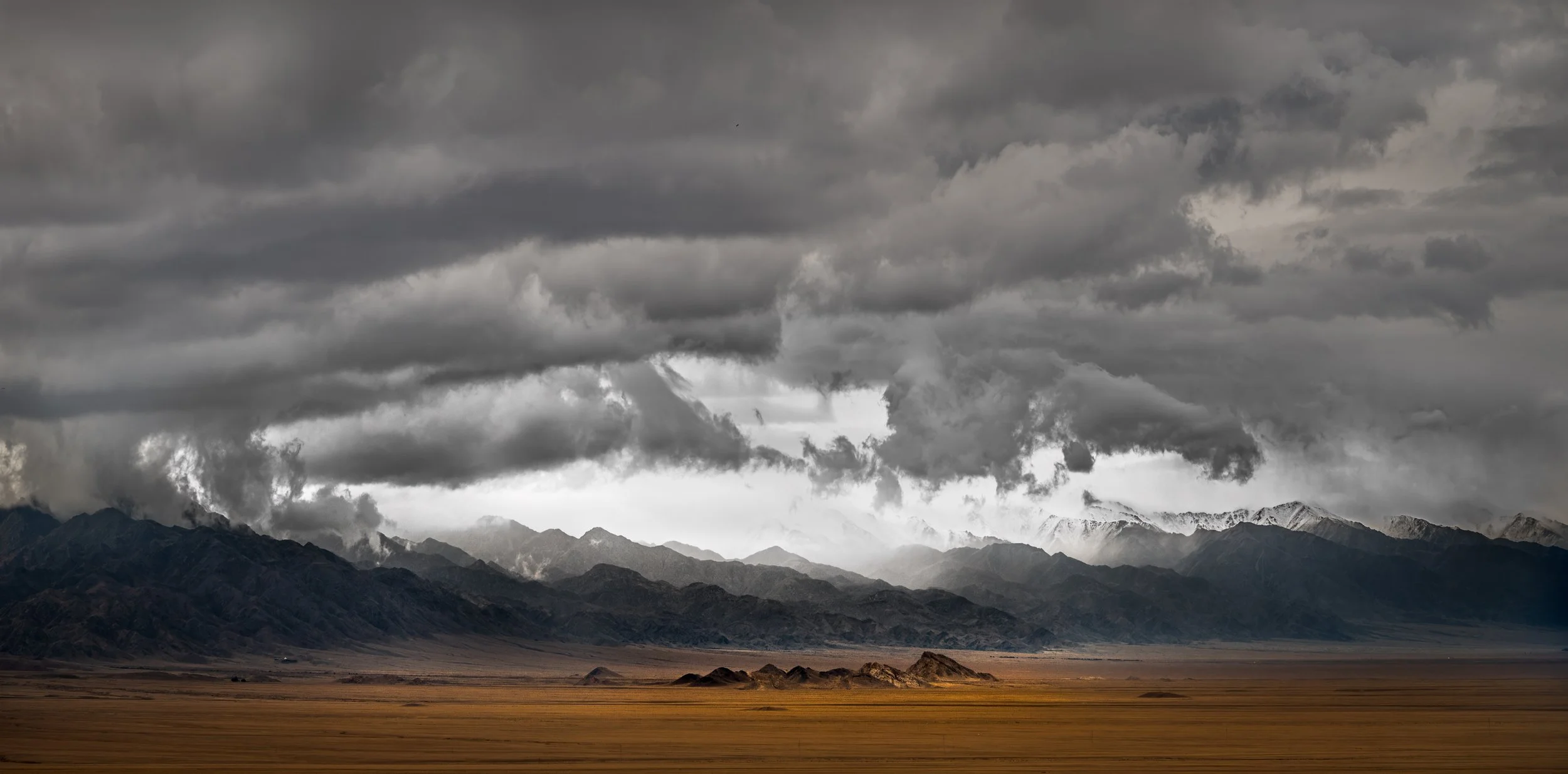 A vast valley with golden grasslands in the foreground, dark rocky mountains in the middle, and snow-capped peaks under a cloudy, stormy sky in the background.