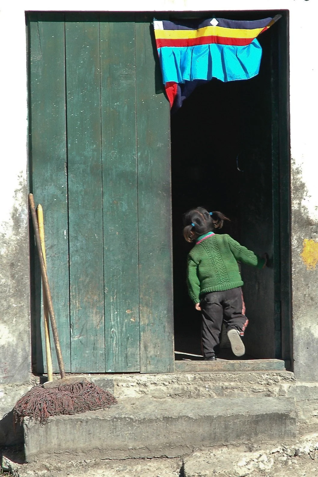 A young girl with pigtails wearing a green sweater and black pants walking into a dark doorway, with a colorful cloth draped above the doorway and a broom resting against the wall outside.