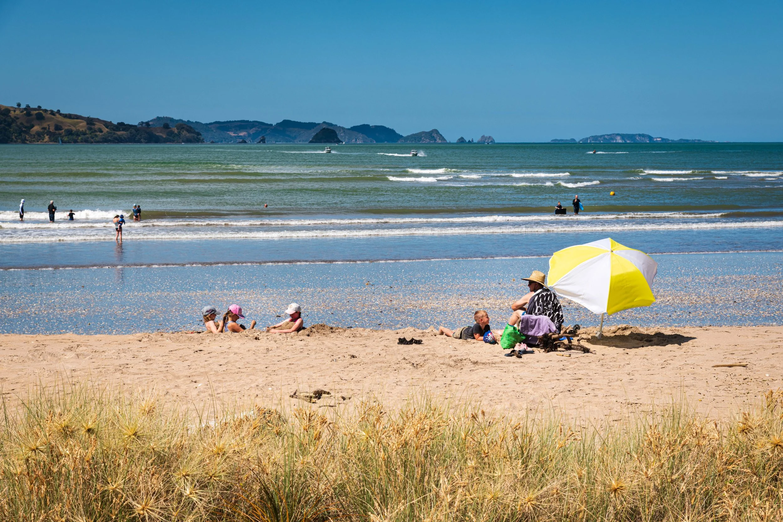 Beach scene with people sitting under a yellow and white umbrella, children playing in the sand, and others in the water, with green hills and small islands in the background.