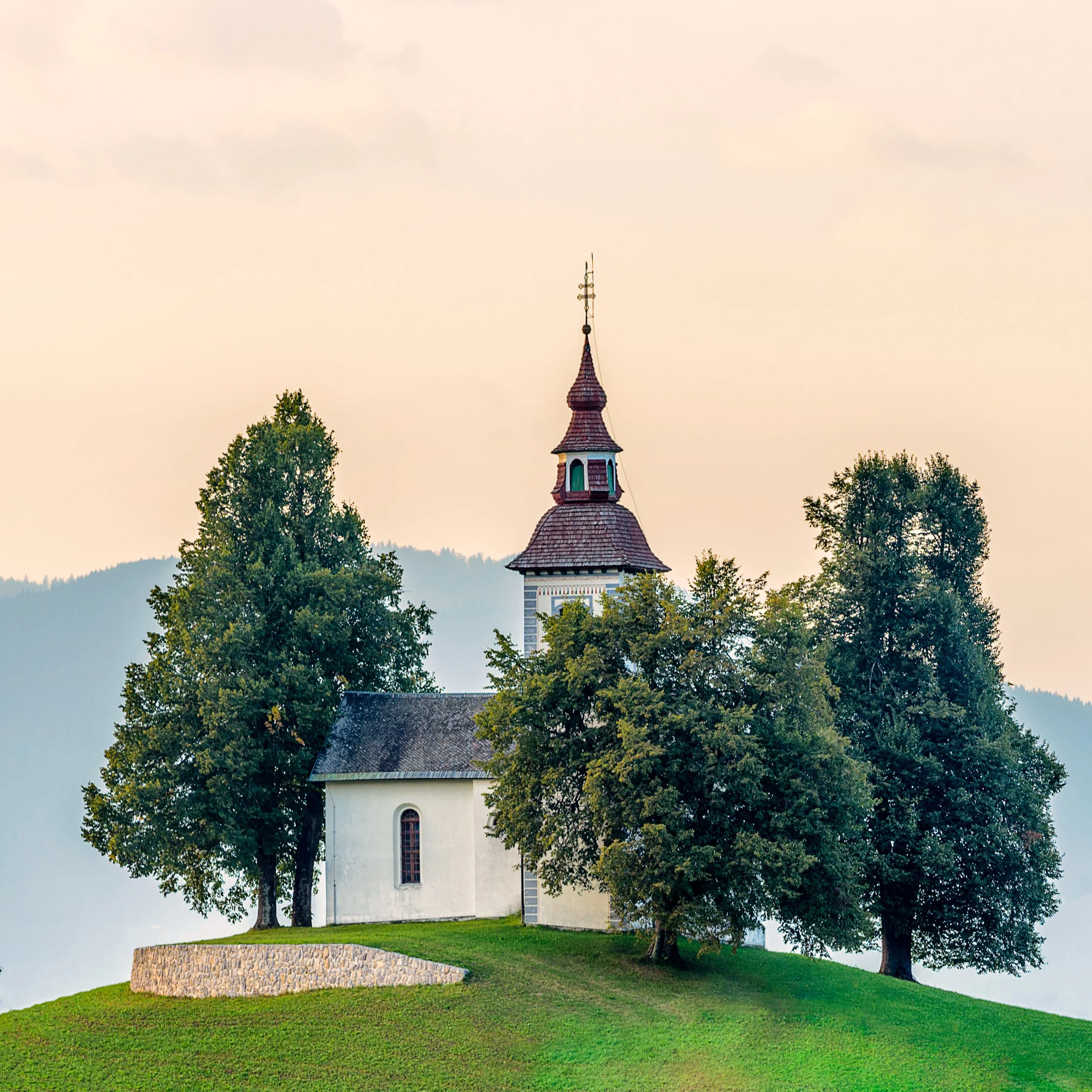 A small white church with a brown roof, surrounded by several green trees on a grassy hill, with mountains in the background under a pale sky.