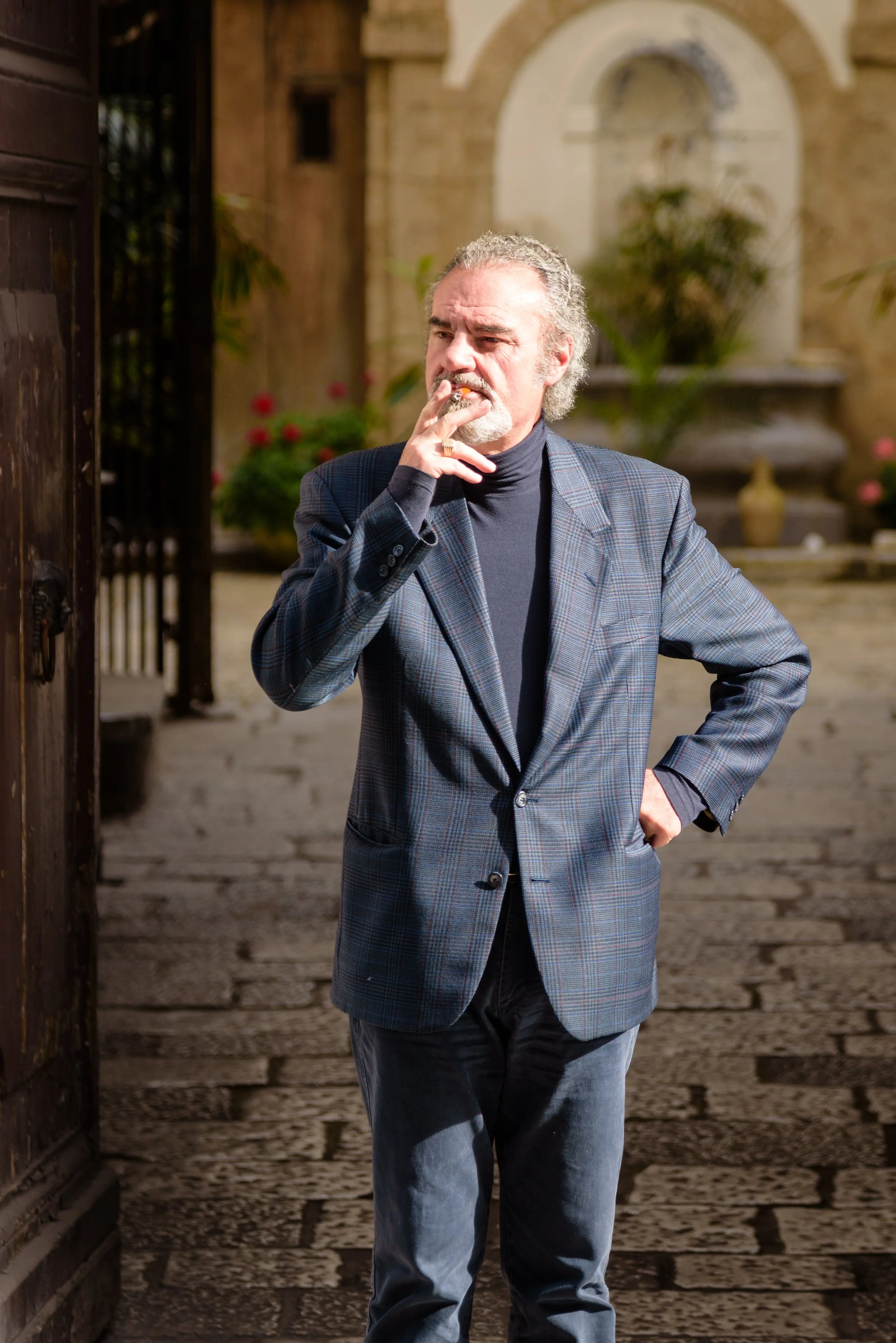 A man with gray hair and a beard, wearing a plaid blazer and black turtleneck, stands outside near a stone fountain, smoking a cigarette.