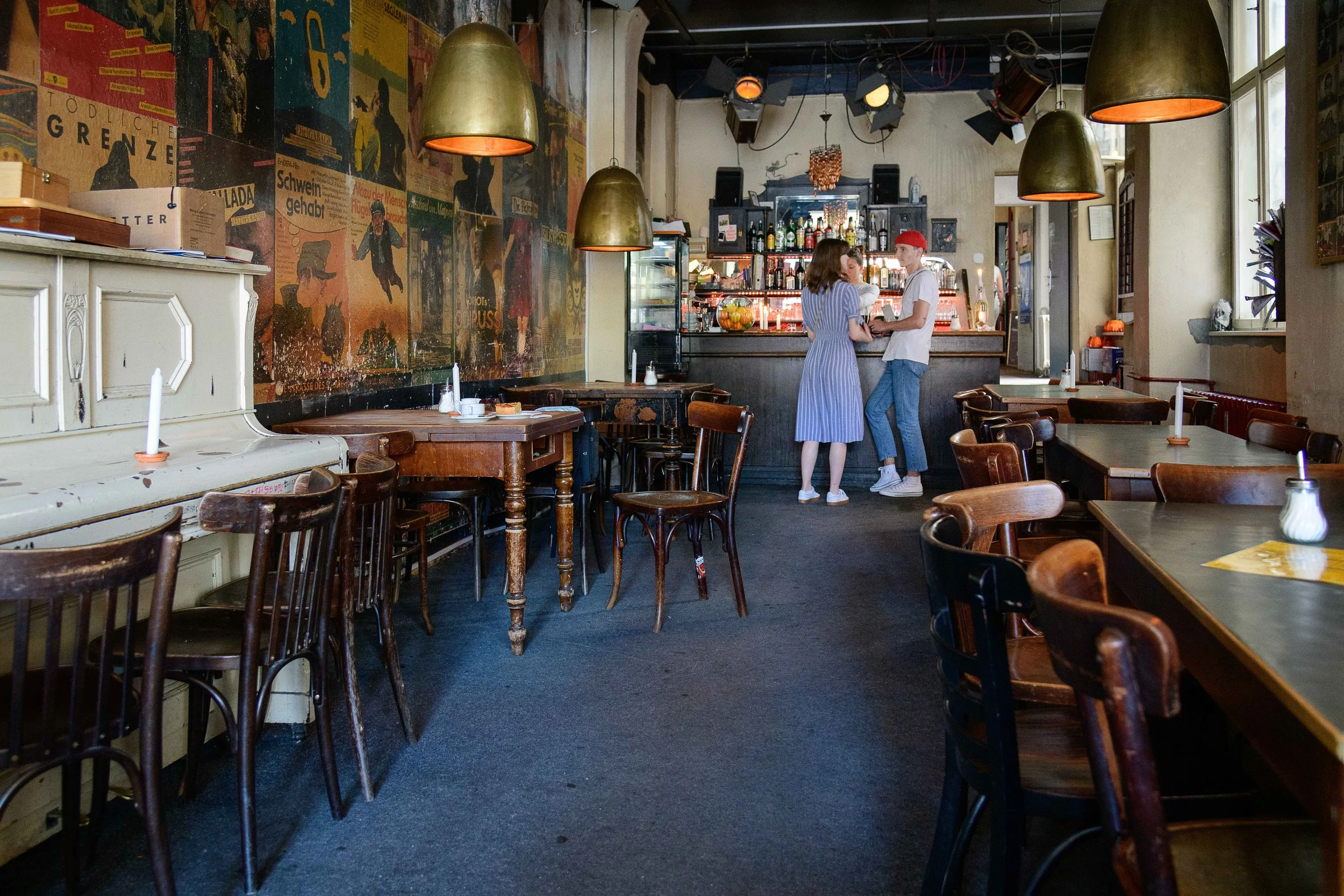 Interior of a cozy vintage-style cafe with wooden tables and chairs, a white piano, and a mural wall. Two people are at the bar, talking, with warm lighting from hanging lamps.