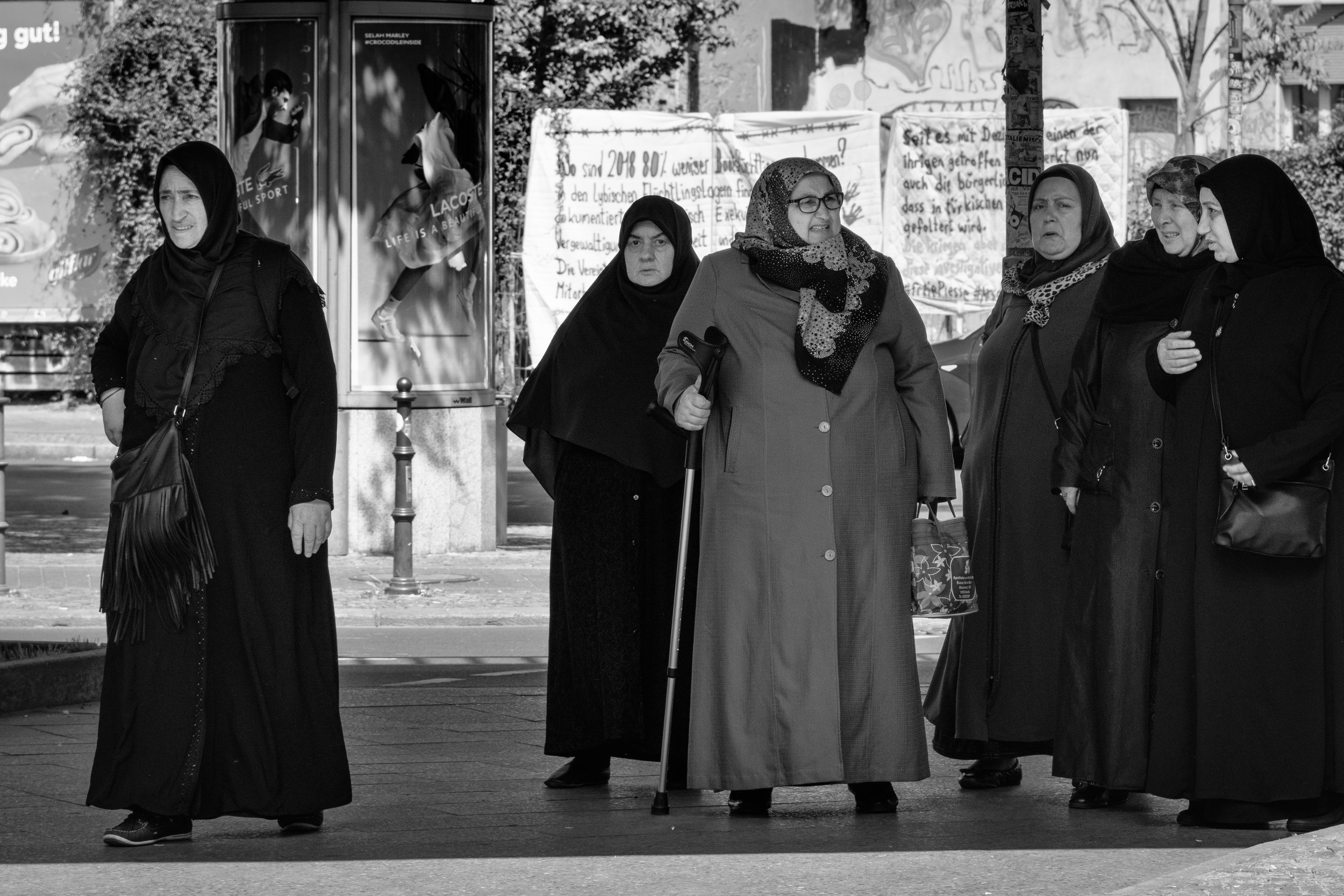 Six women wearing hijabs and long coats standing on a city sidewalk, some engaged in conversation, in black and white.