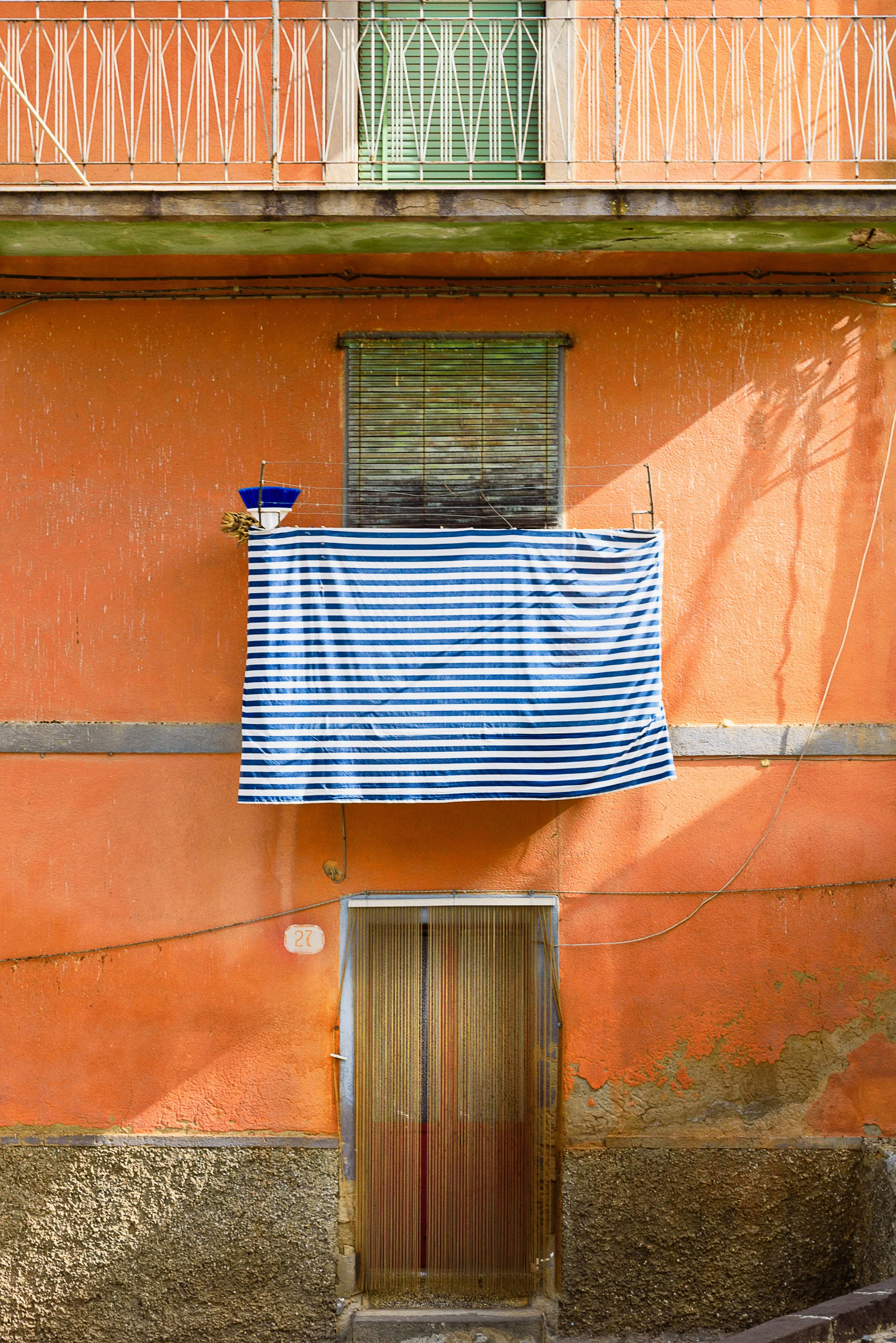 Orange building with a small balcony, a striped blue and white fabric hanging on a clothesline, a window with closed blinds, and a door with a translucent covering.