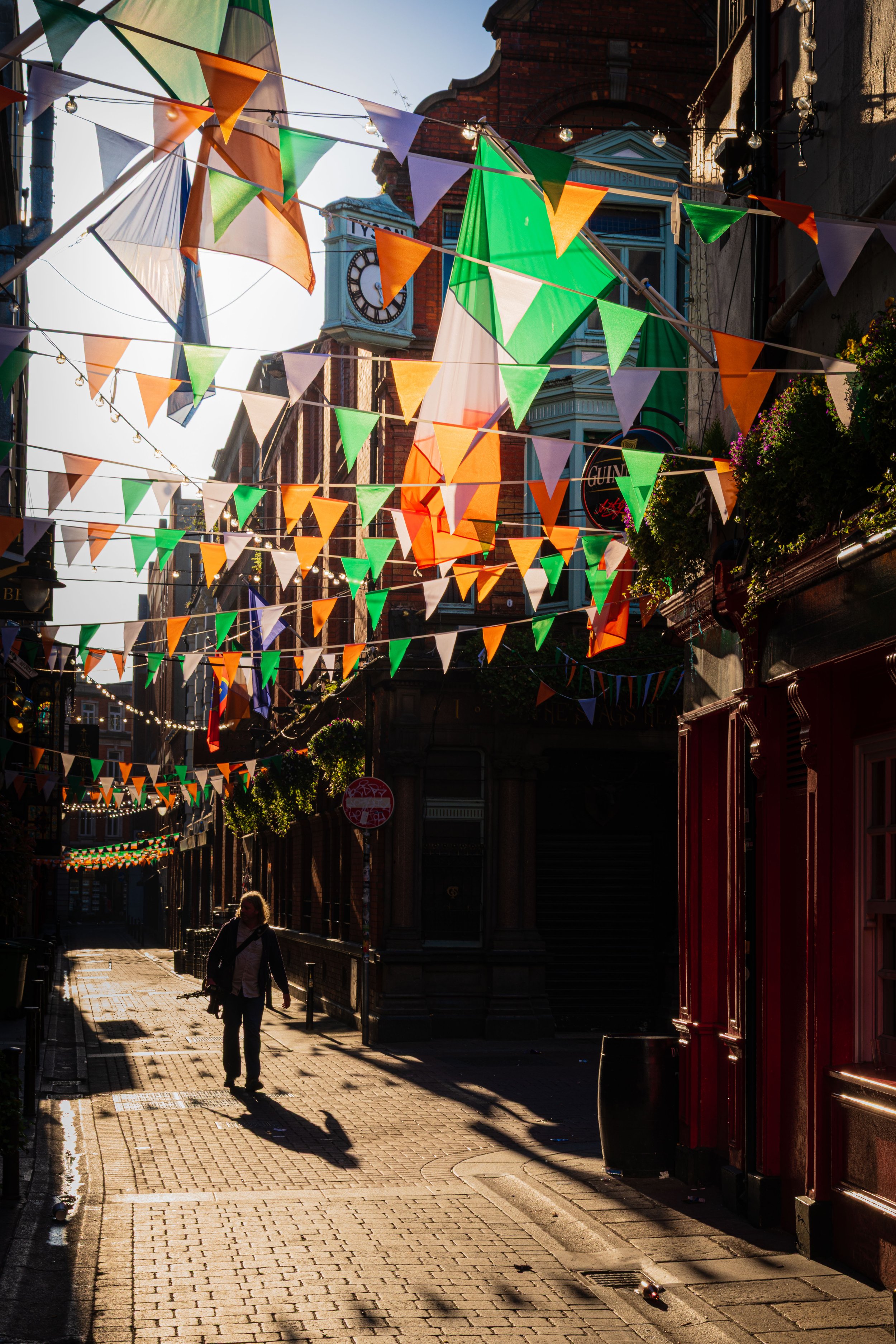 A person walking down a narrow street decorated with colorful triangular flags overhead, with buildings and a clock tower in the background, during sunset.