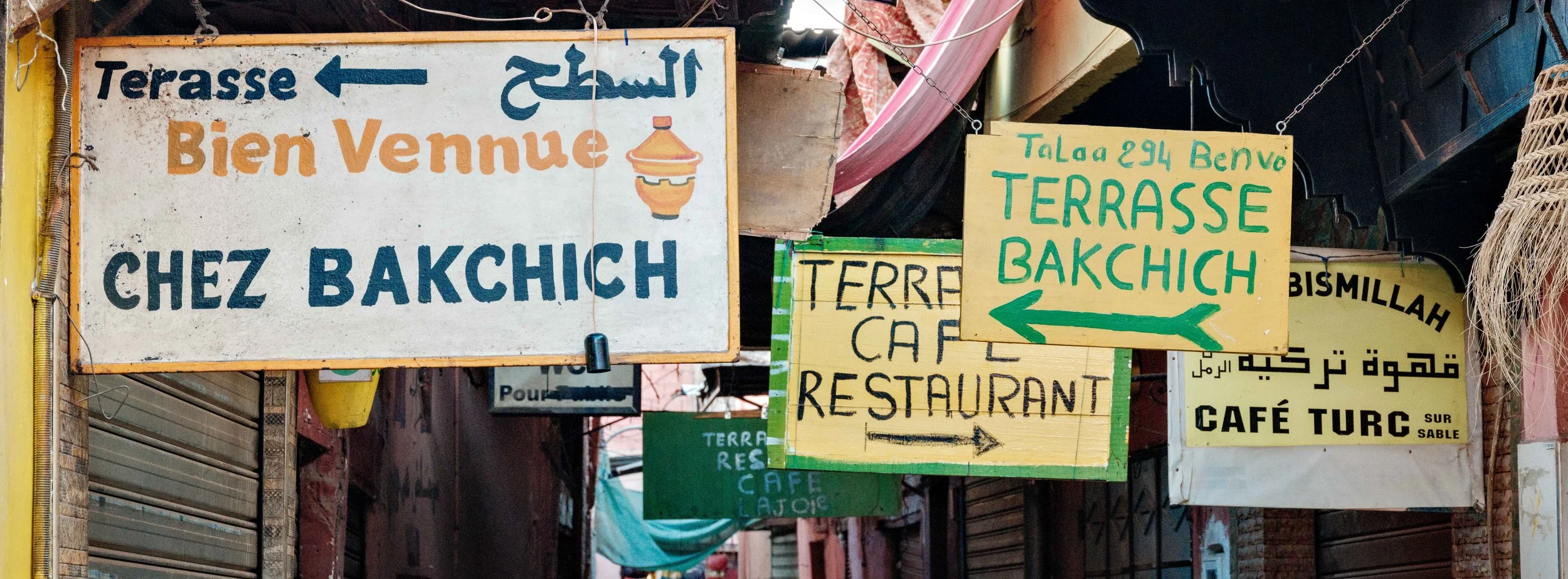 Several colorful signs in multiple languages hanging above a street, welcoming visitors to a terrace restaurant and directing to cafes and other local attractions.