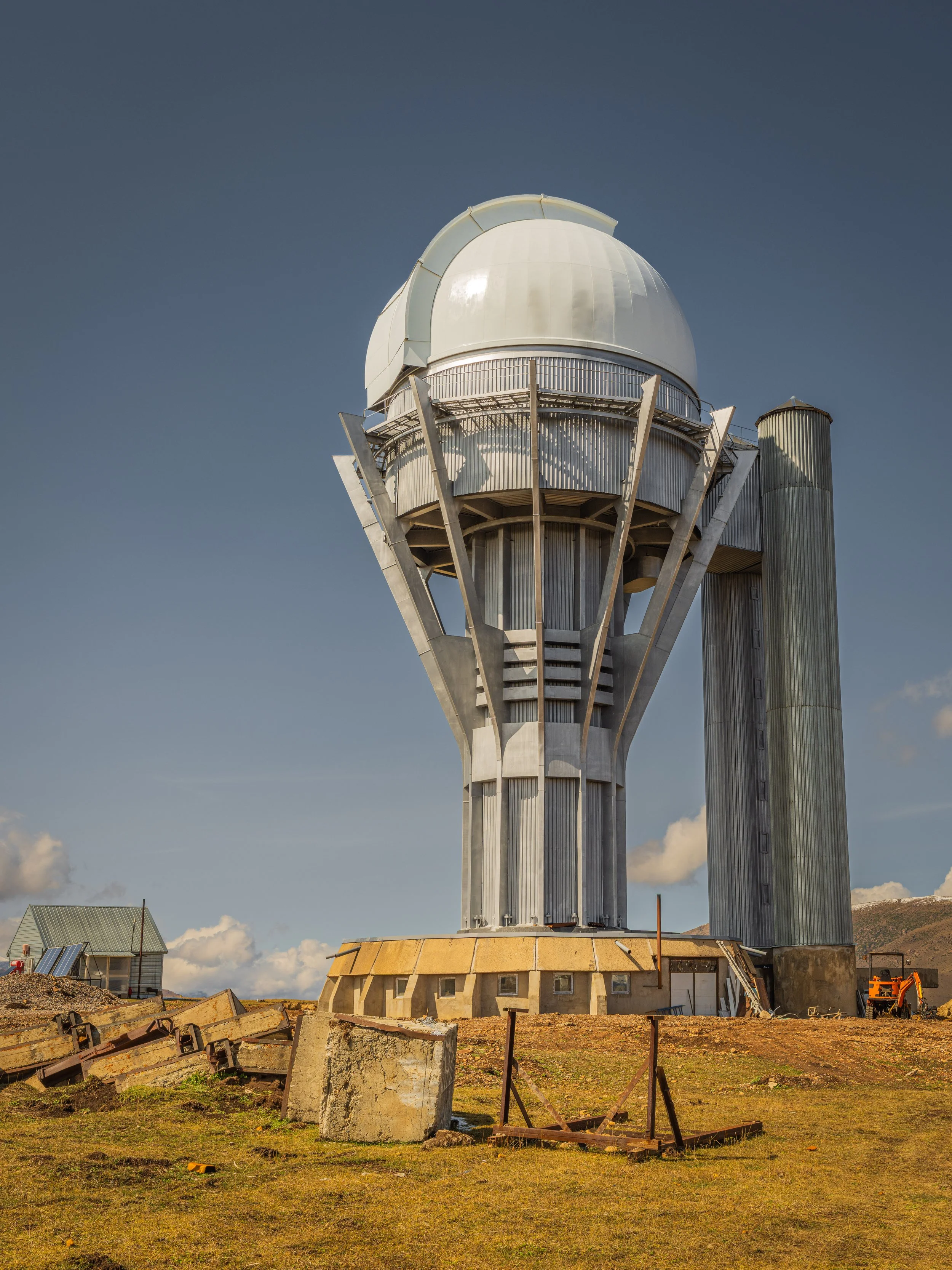 A large observatory with a white dome and tall supporting structure against a clear sky, situated on a grassy plain with some scattered rocks.