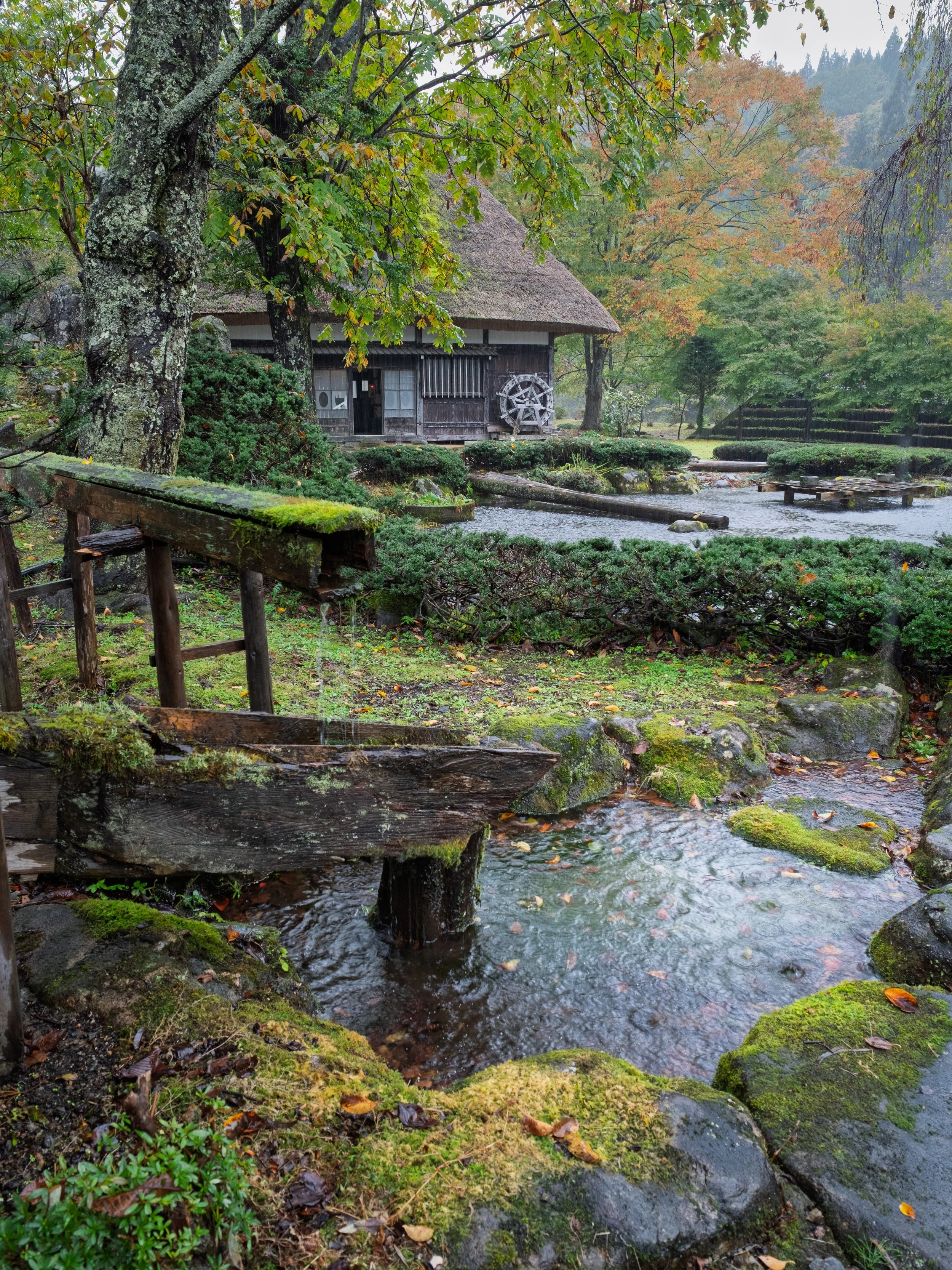A traditional Japanese garden with a small stream, moss-covered rocks, wooden footbridge, lush green and autumn-colored trees, and a thatched-roof house with a water wheel, creating a peaceful, natural scene.