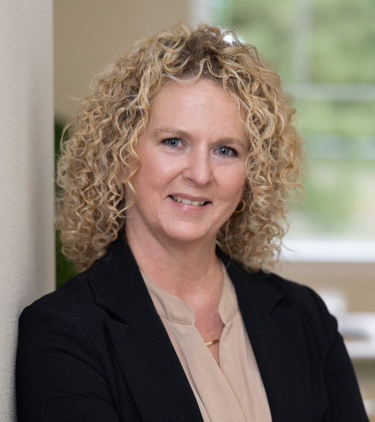 A woman with curly blonde hair wearing a black blazer and a beige blouse, standing indoors near a wall with a window showing green trees outside.