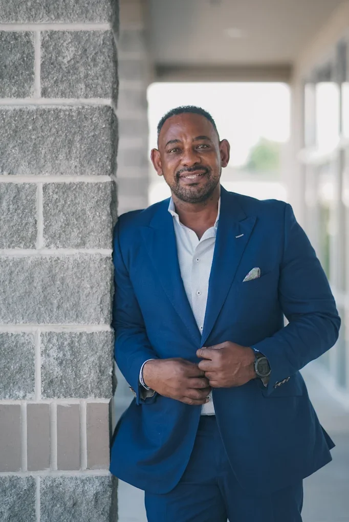 A man in a blue suit leaning against a brick wall outside a building, smiling.