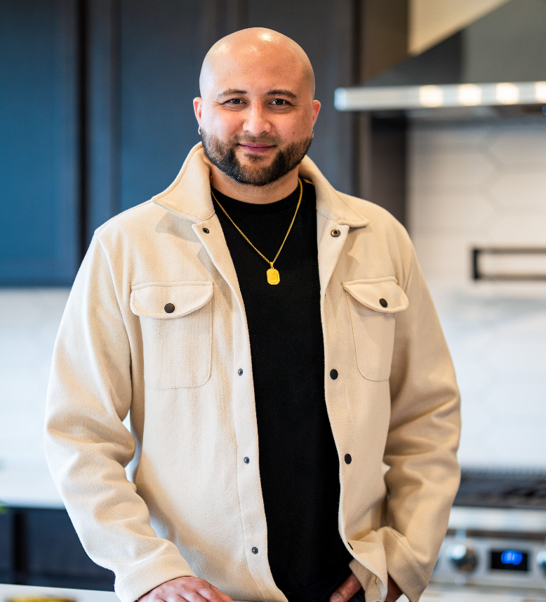 A man with a shaved head and beard standing in a modern kitchen, wearing a beige jacket over a black shirt and a gold necklace.