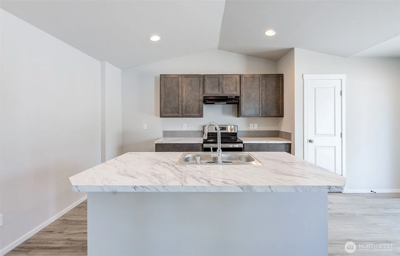 A modern kitchen with white walls, a marble countertop island with a sink, and dark wood cabinets, with wood flooring and recessed ceiling lights.