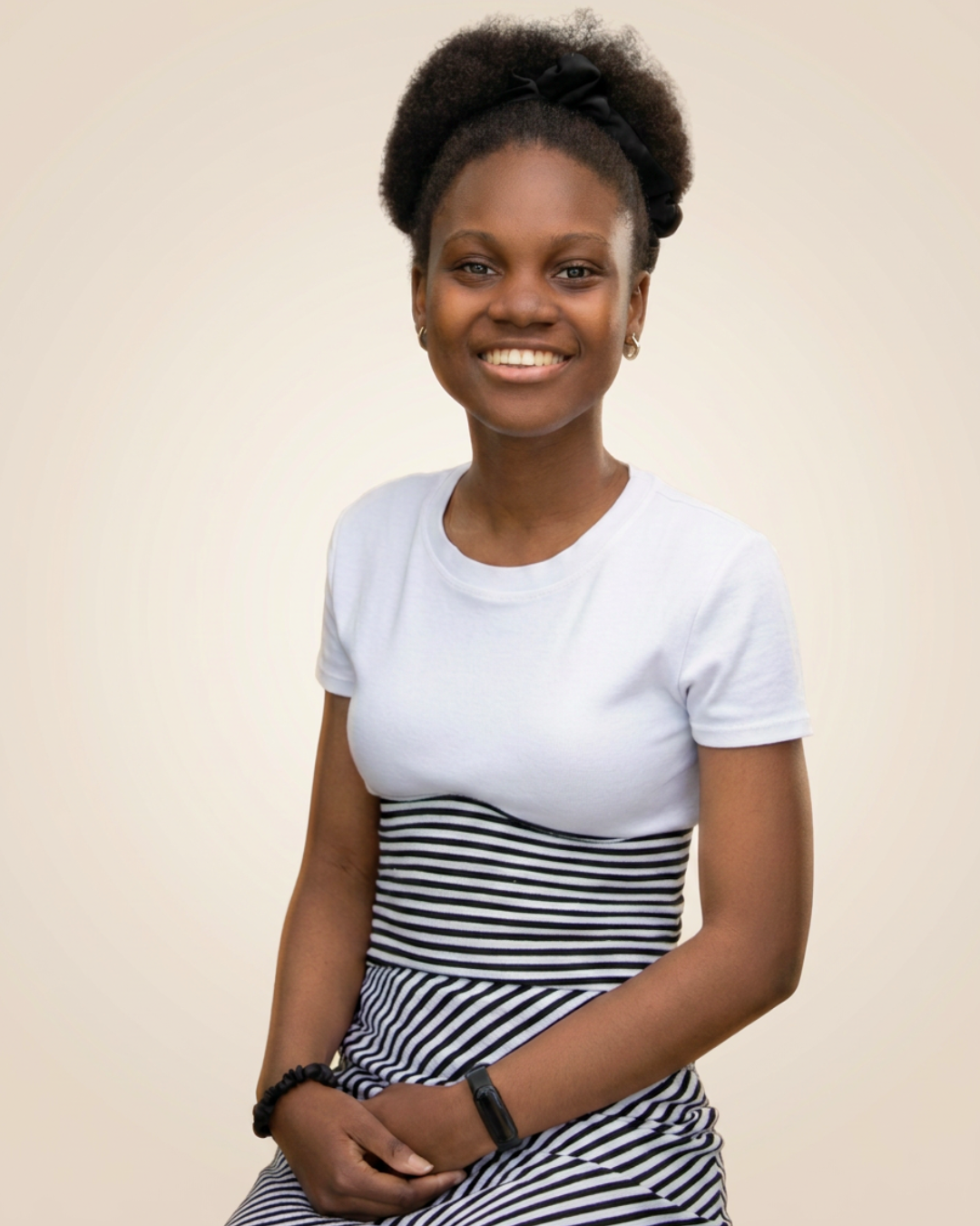 A young woman with a big smile, wearing a white t-shirt and a black and white striped skirt, sitting against a light-colored background.