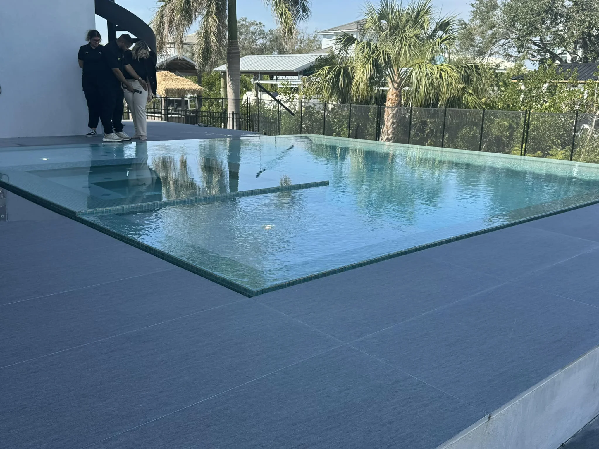 Four people stand on the deck of a swimming pool, looking down. The pool is outdoors with a glass wall and palm trees in the background, under a blue sky.