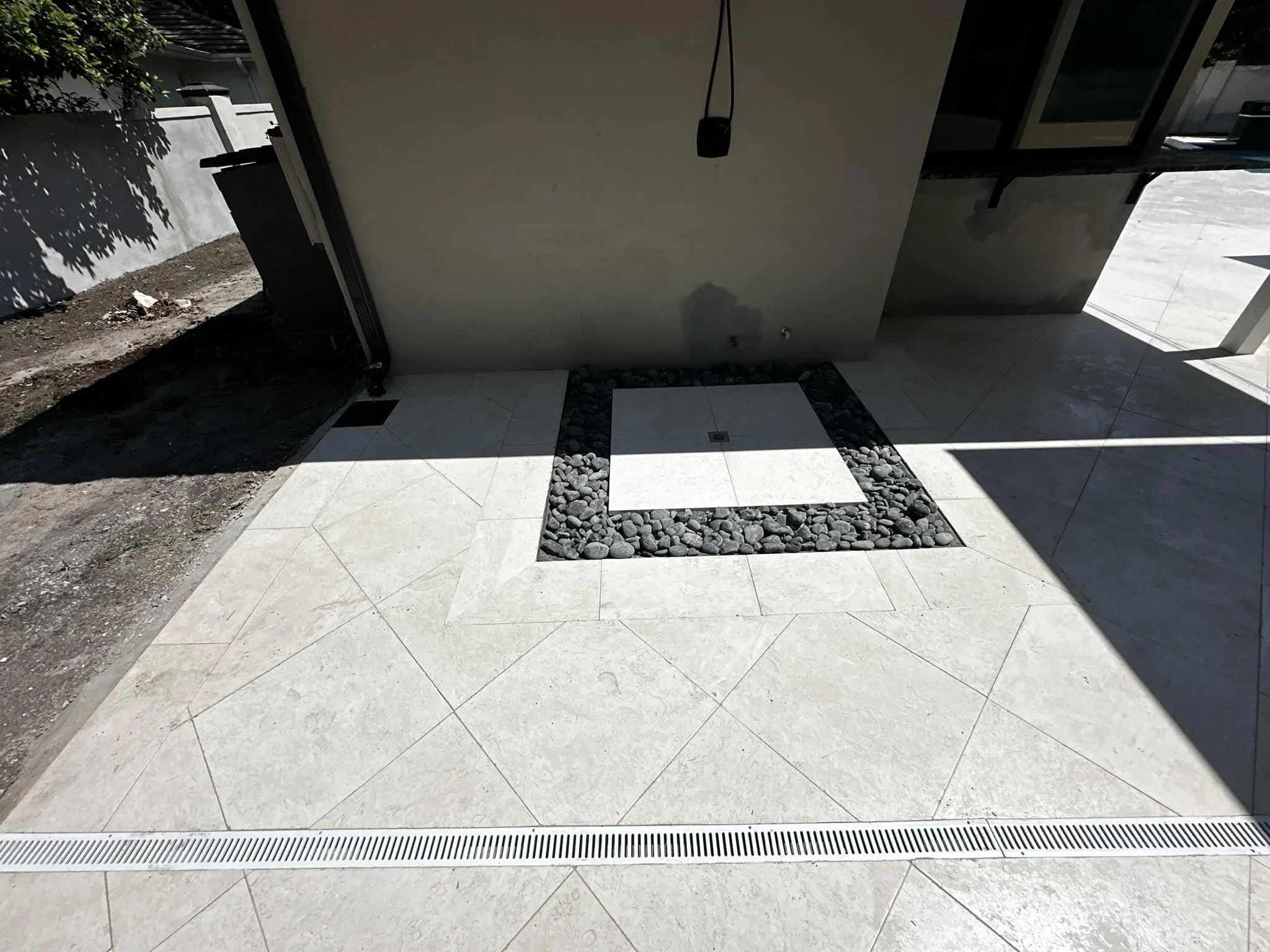 View of an outdoor shower area with a tiled floor, a square drain, and a wall with a shower head and hose. Part of a building and some dirt ground are visible.