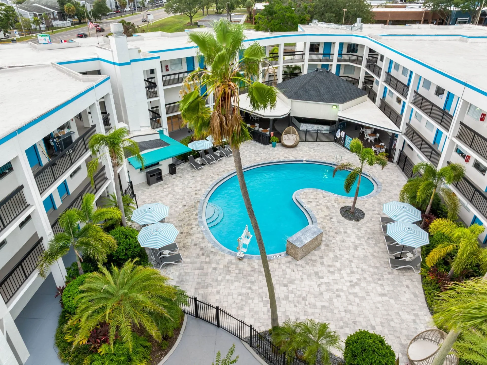 An aerial view of a hotel courtyard featuring a swimming pool surrounded by patio chairs with umbrellas, palm trees, and a multi-story building with balconies.