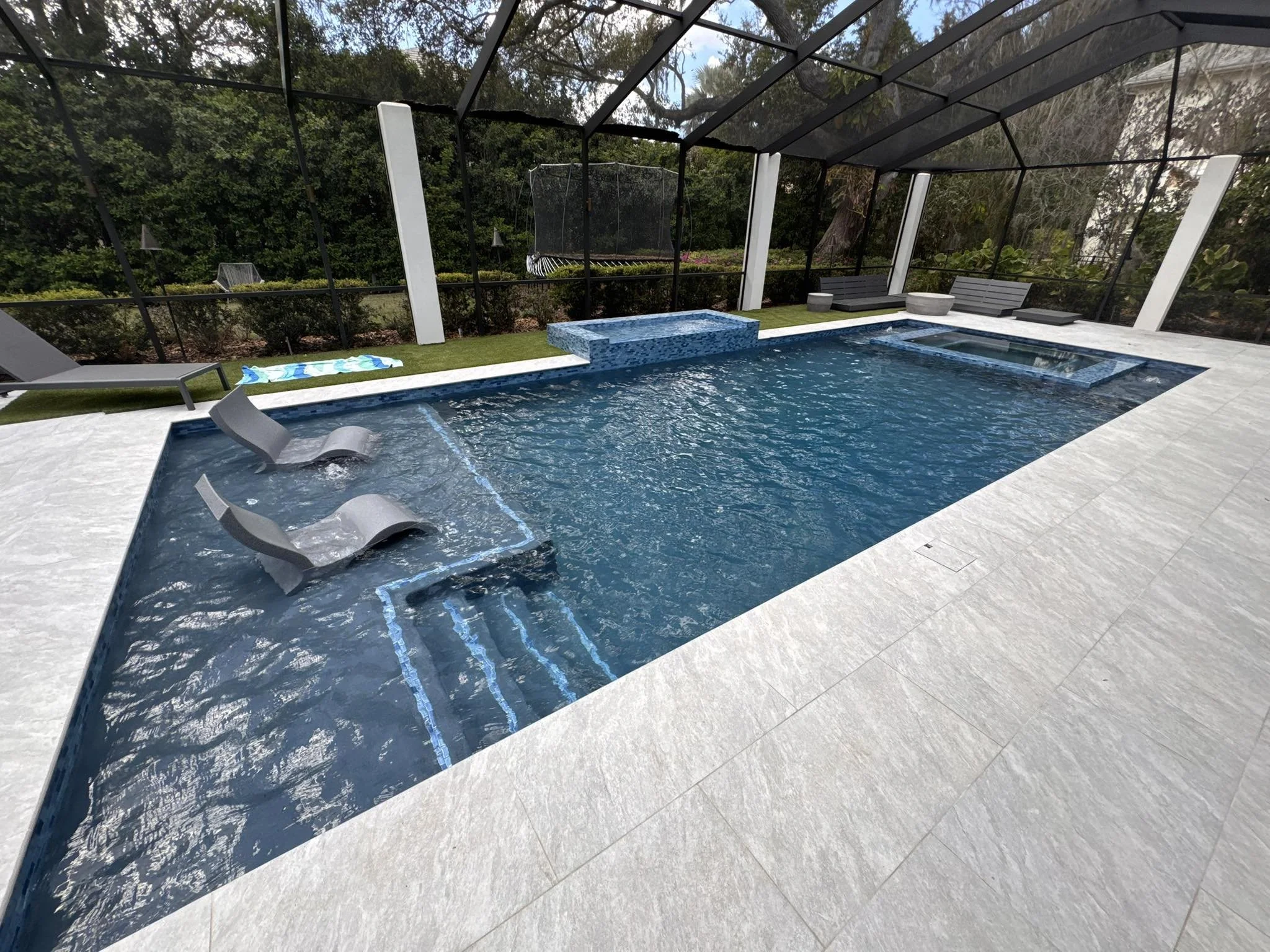 A modern backyard pool area enclosed by a glass screen with a mesh net. The pool features built-in lounge chairs submerged in water, a spa section, and a shallow seating area with steps. There are lounge chairs, a towel, and outdoor seating around the pool. Trees and greenery are visible outside the enclosure.