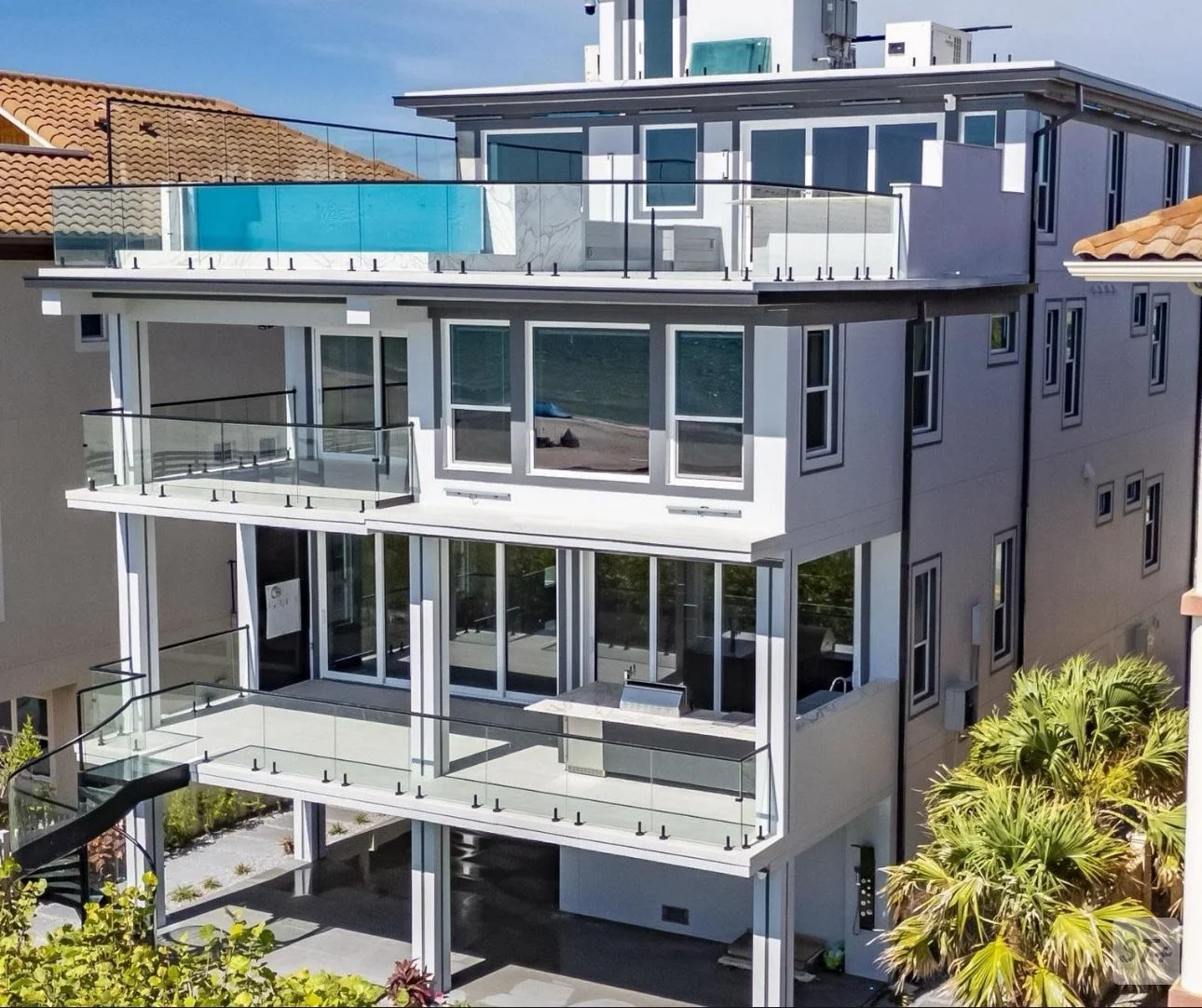 Modern multi-story residential building with glass balconies, large windows, and a rooftop pool with a glass safety barrier. Surrounding palm trees and neighboring houses are visible.