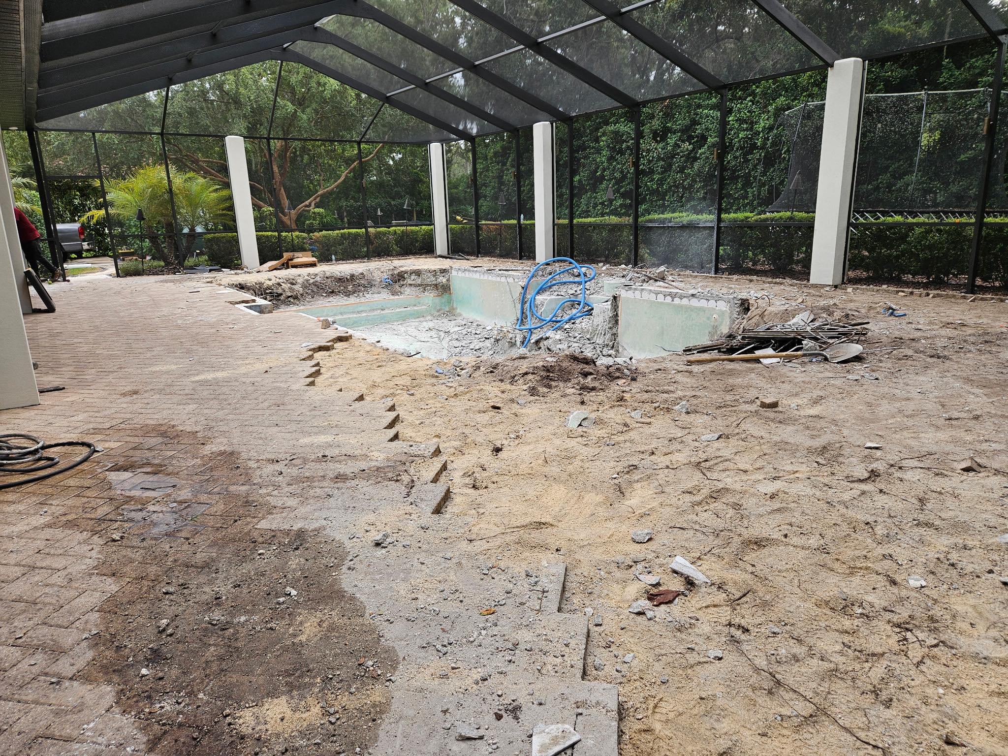 Empty swimming pool under renovation with construction debris, exposed wiring, and partially removed bricks inside a screened backyard area surrounded by trees and bushes.