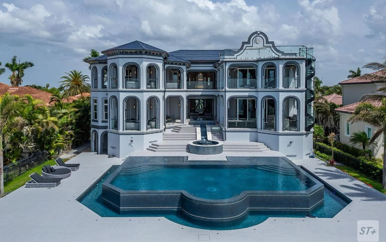 Large luxurious white mansion with multiple levels and arched windows, surrounded by palm trees and other tropical plants, with a large in-ground swimming pool and hot tub in the backyard.