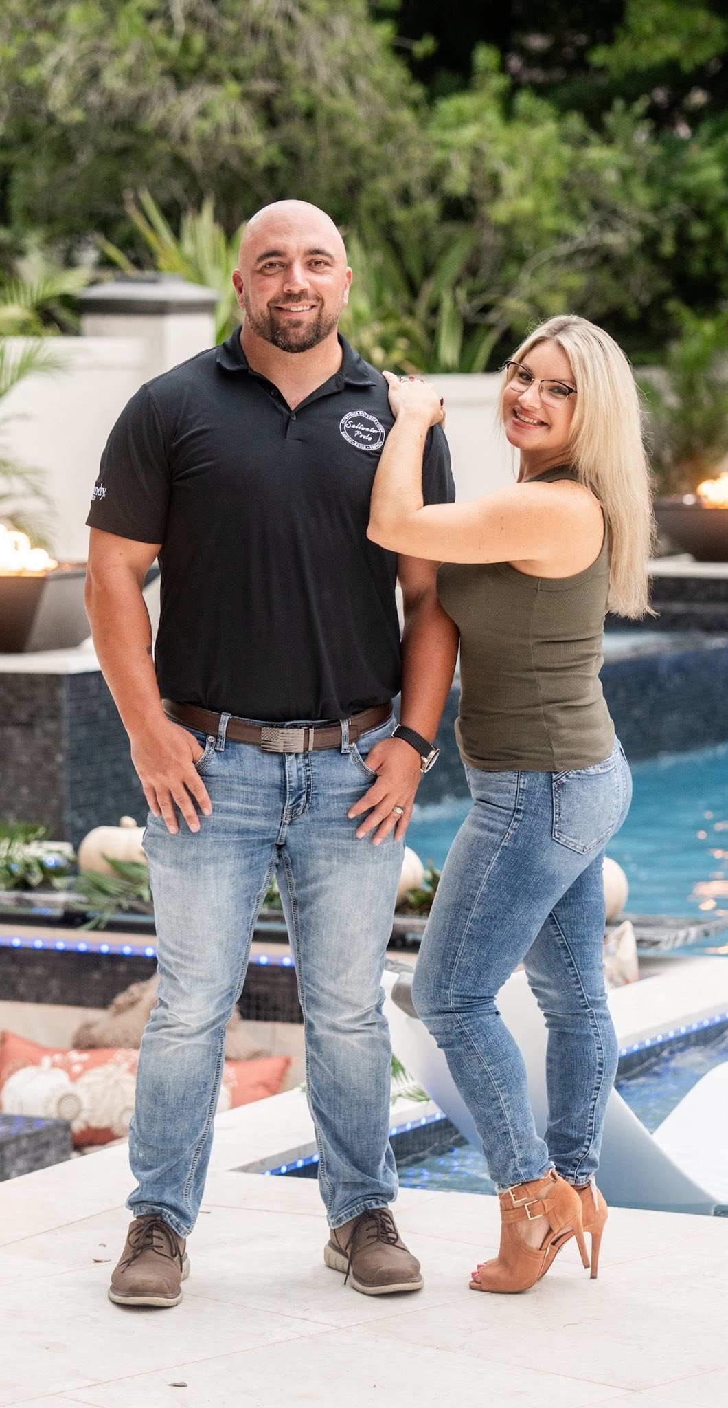 A man and woman standing outdoors by a pool at sunset, smiling, with the woman touching the man's shoulder.