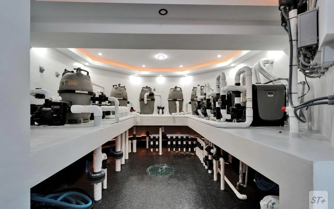 Indoor pool pump room with white plumbing pipes, filters, and equipment arranged on white counters, under ceiling lights and a false ceiling with orange strip lighting.