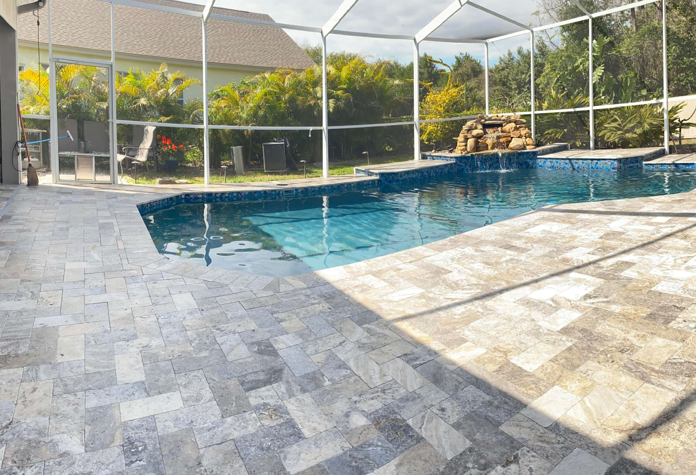 A backyard swimming pool enclosed by a screened patio with a waterfall feature, surrounded by a stone-tiled deck and lush greenery outside.