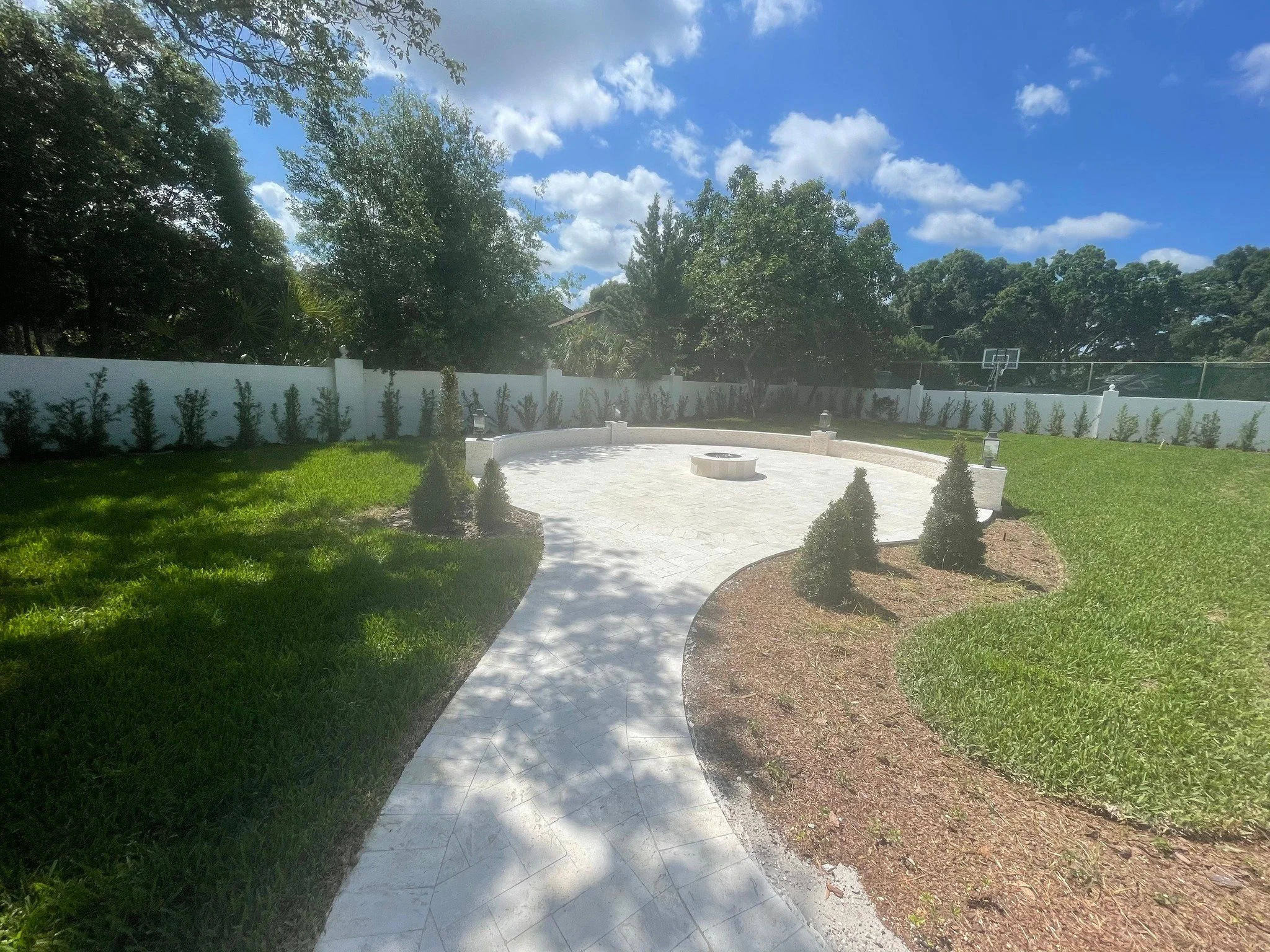 A backyard with a curved white stone pathway leading to a wheelchair basketball area, surrounded by a white fence and lush green trees under a partly cloudy blue sky.