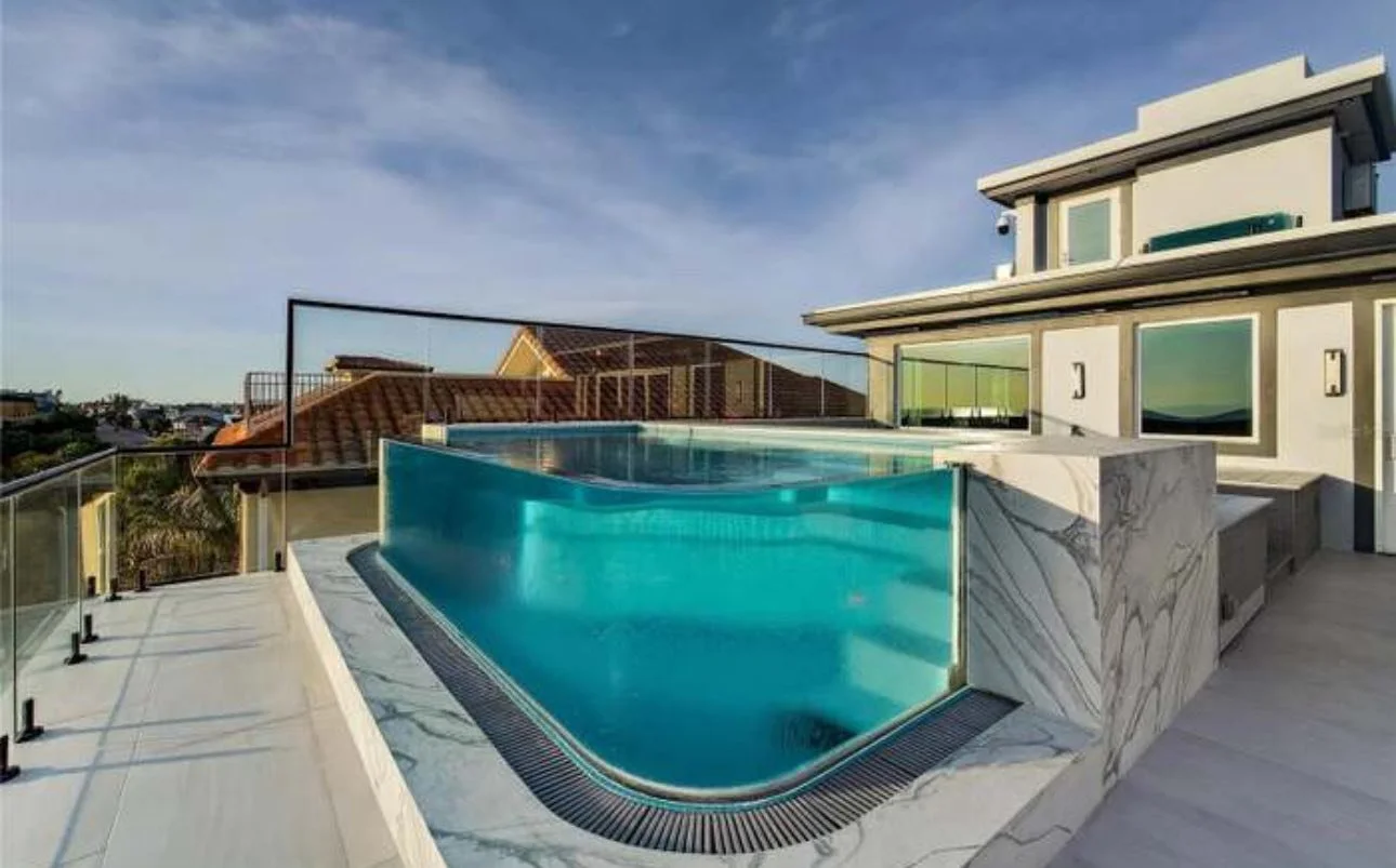Modern rooftop infinity pool with glass fencing surrounding it, overlooking a neighborhood with houses and a clear sky.