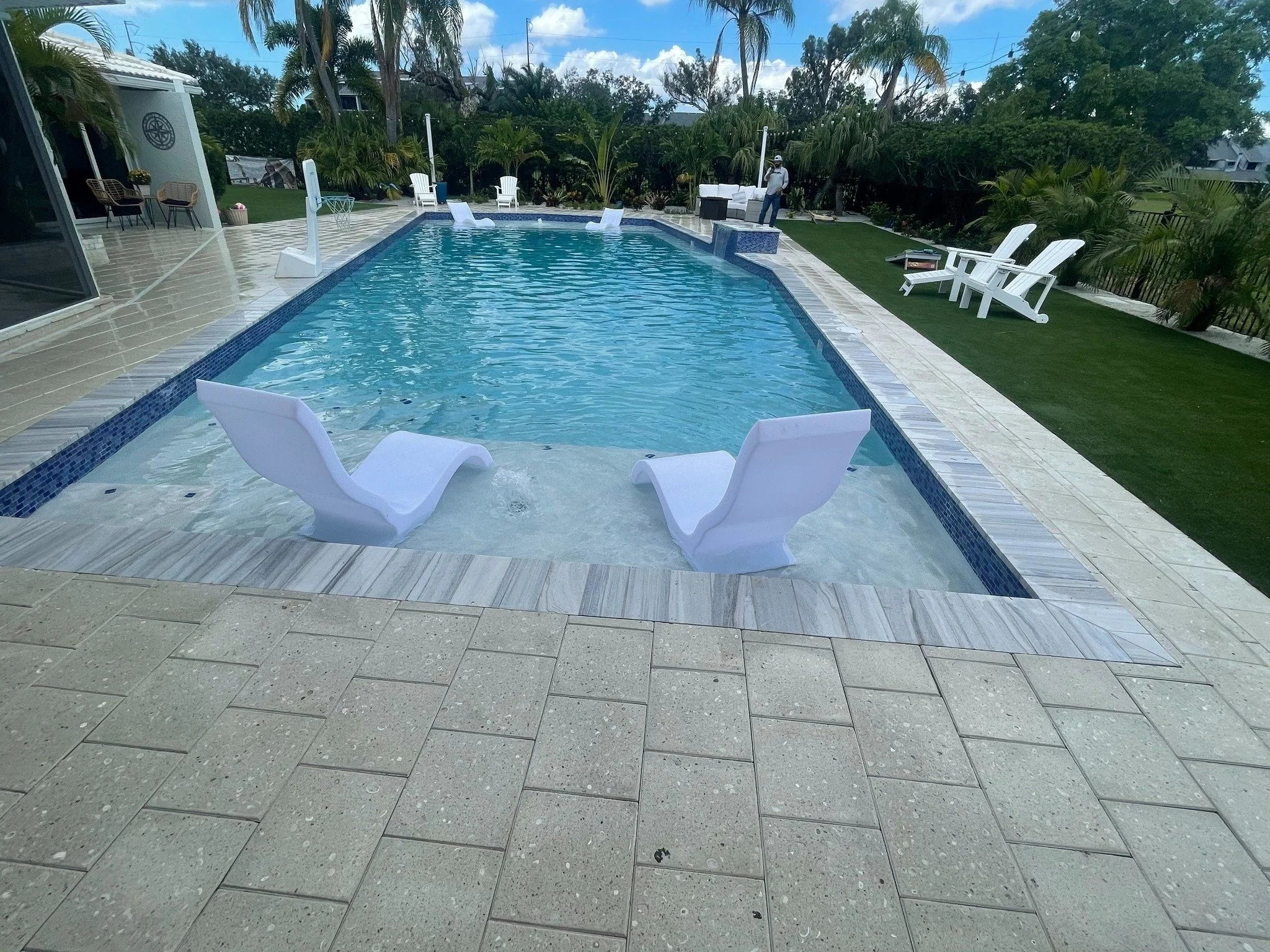 A backyard swimming pool with two modern, white lounge chairs partially submerged in the water at the shallow end. The pool is surrounded by a tiled patio, palm trees, and lawn chairs on a grassy area. A person stands near the deep end of the pool, and there are various outdoor furniture pieces and lush greenery in the background.