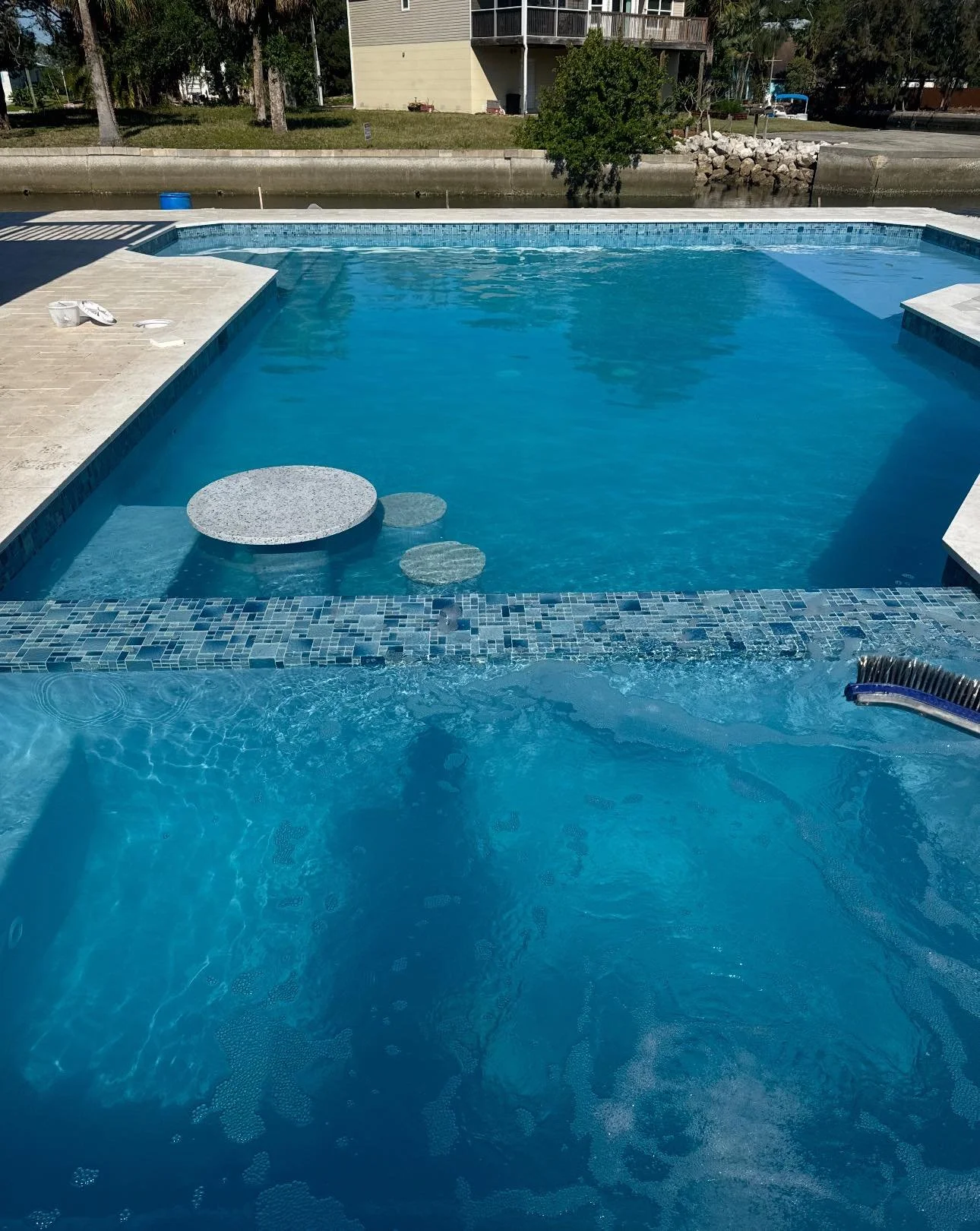 Swimming pool with pool toys, surrounded by a paved deck, overlooking a canal with trees and buildings in the background.