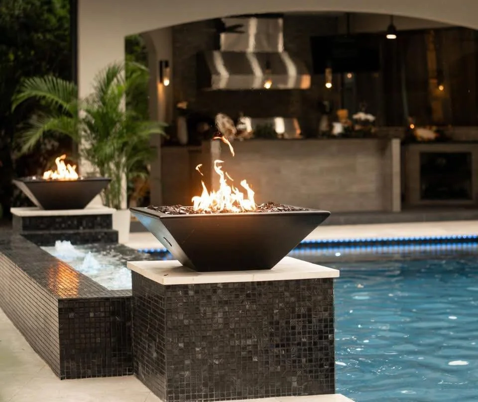 Two modern fire pits with flames placed on black tiled pillars along a swimming pool at night.