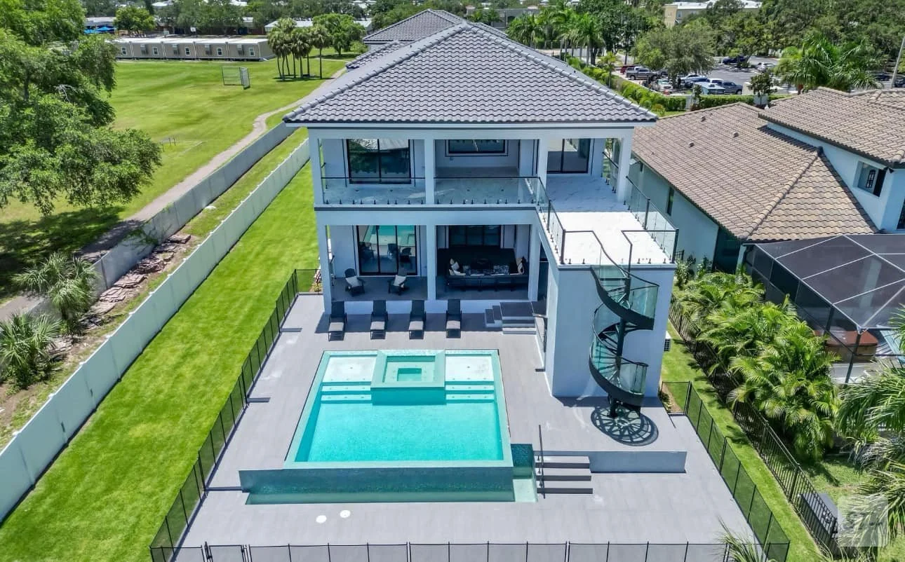 Aerial view of a modern two-story house with a swimming pool, spiral slide, and outdoor seating area in the backyard, surrounded by a fence and nearby houses and greenery.